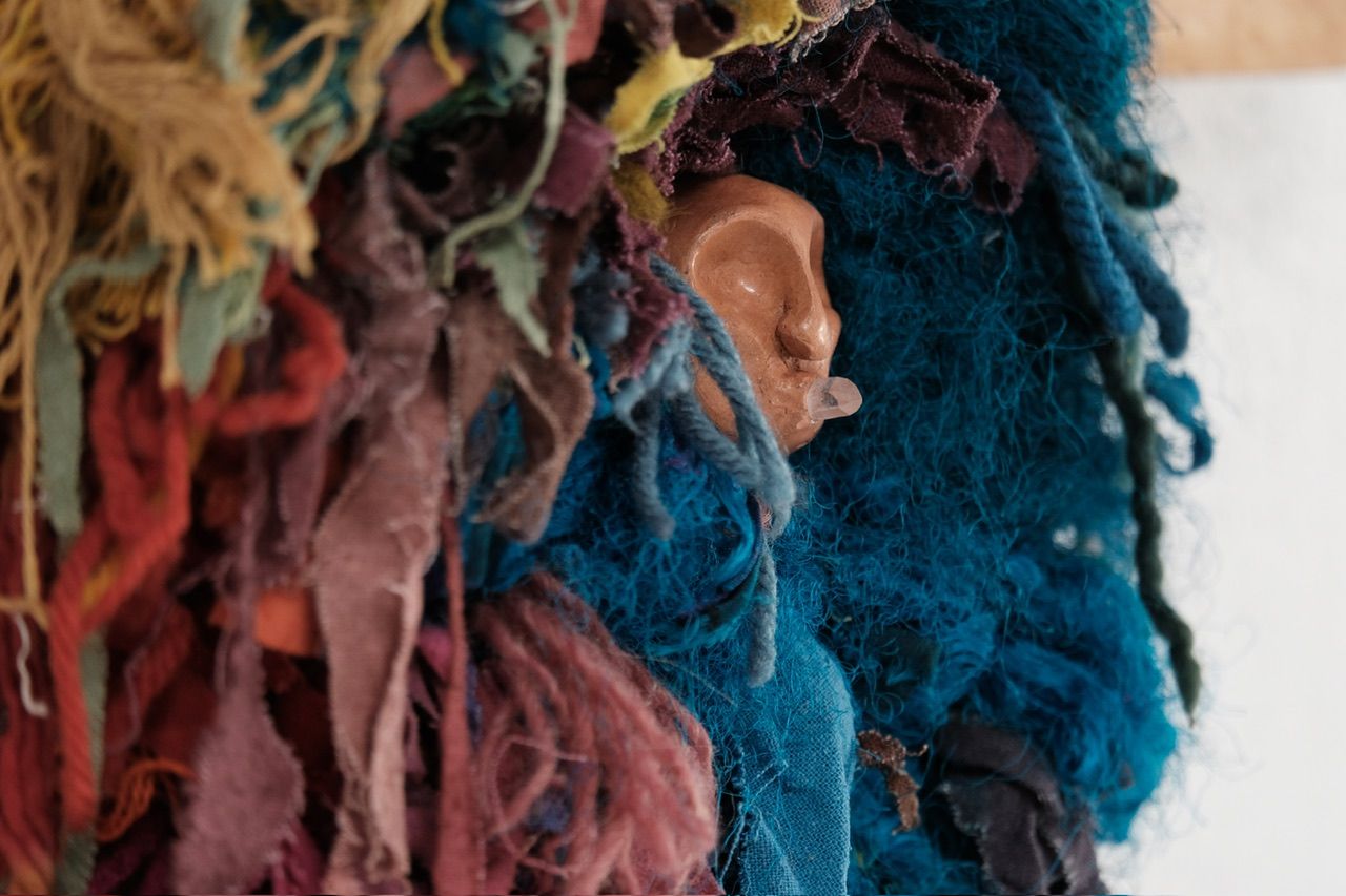 A fiber art sculpture made of yarn and scraps of fabric in a rainbow of colors with a clay face nestled into it, hanging from a maple wood frame on a white wall