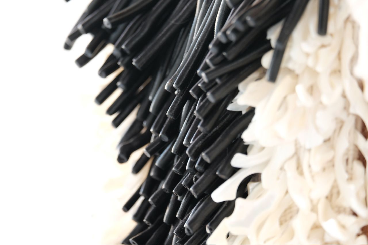 A handwoven sculptural necklace of black coral and mother of pearl hanging on a wooden frame On a white wall