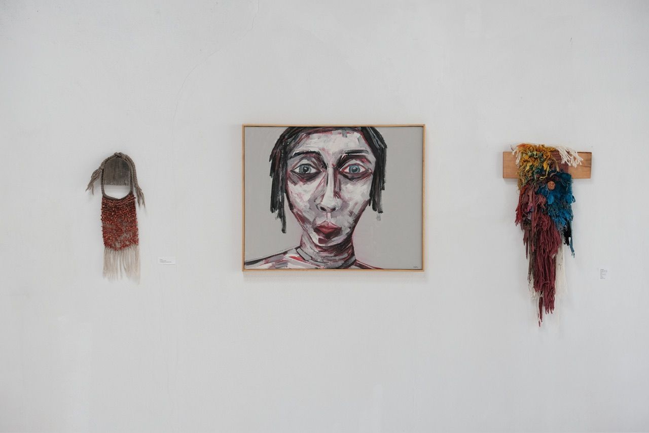 A fiber art sculpture made of yarn and scraps of fabric in a rainbow of colors with a clay face nestled into it, hanging from a maple wood frame on a white wall