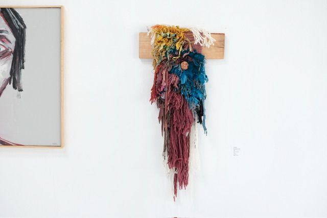 A fiber art sculpture made of yarn and scraps of fabric in a rainbow of colors with a clay face nestled into it, hanging from a maple wood frame on a white wall