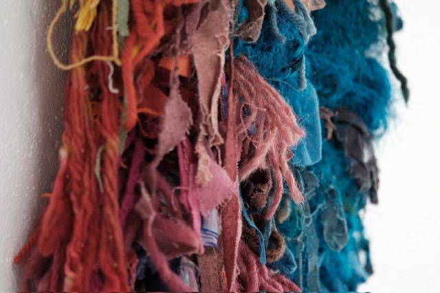 A fiber art sculpture made of yarn and scraps of fabric in a rainbow of colors with a clay face nestled into it, hanging from a maple wood frame on a white wall