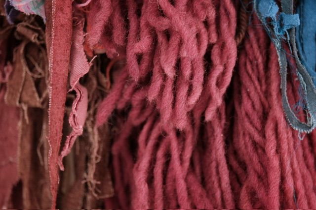 A fiber art sculpture made of yarn and scraps of fabric in a rainbow of colors with a clay face nestled into it, hanging from a maple wood frame on a white wall