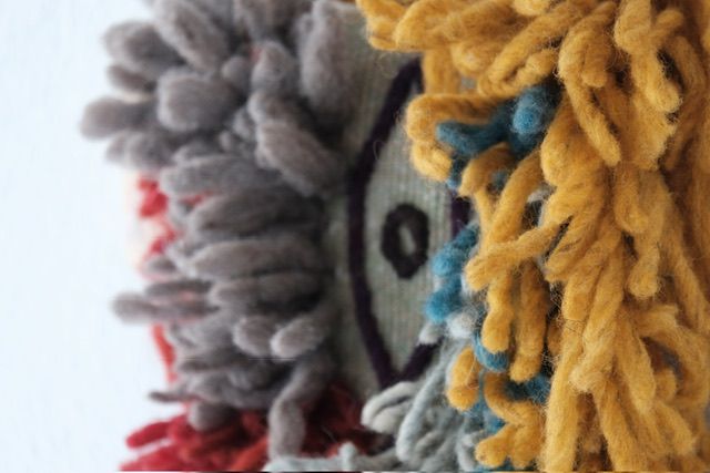 Handwoven many-colored pile woven churro wool wall hanging sculpture with a clay face emerging from it with purple yarn coming from its mouth