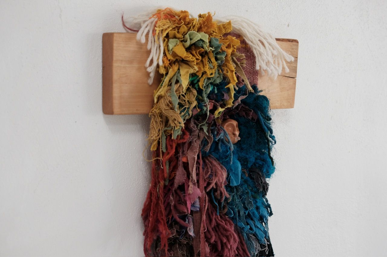 A fiber art sculpture made of yarn and scraps of fabric in a rainbow of colors with a clay face nestled into it, hanging from a maple wood frame on a white wall