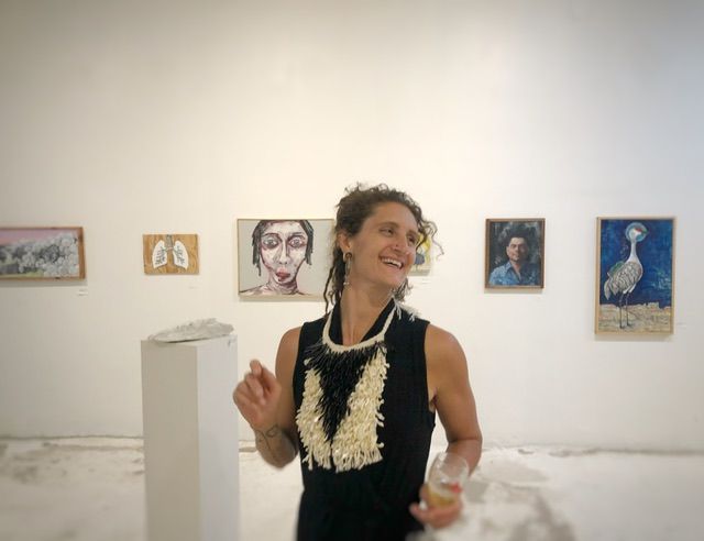 A woman is wearing a black outfit and a sculptural mother of pearl and black coral necklace stands against a gallery wall with two oil paintings on each side