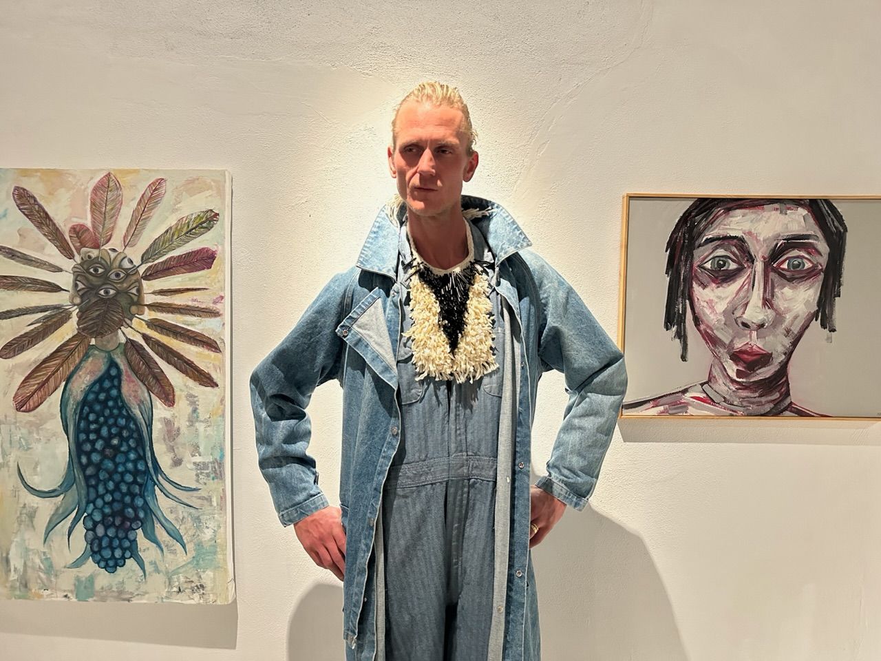 A tall thin man wearing a light denim outfit and a sculptural mother of pearl and black coral necklace stands against a gallery wall with two oil paintings on each side