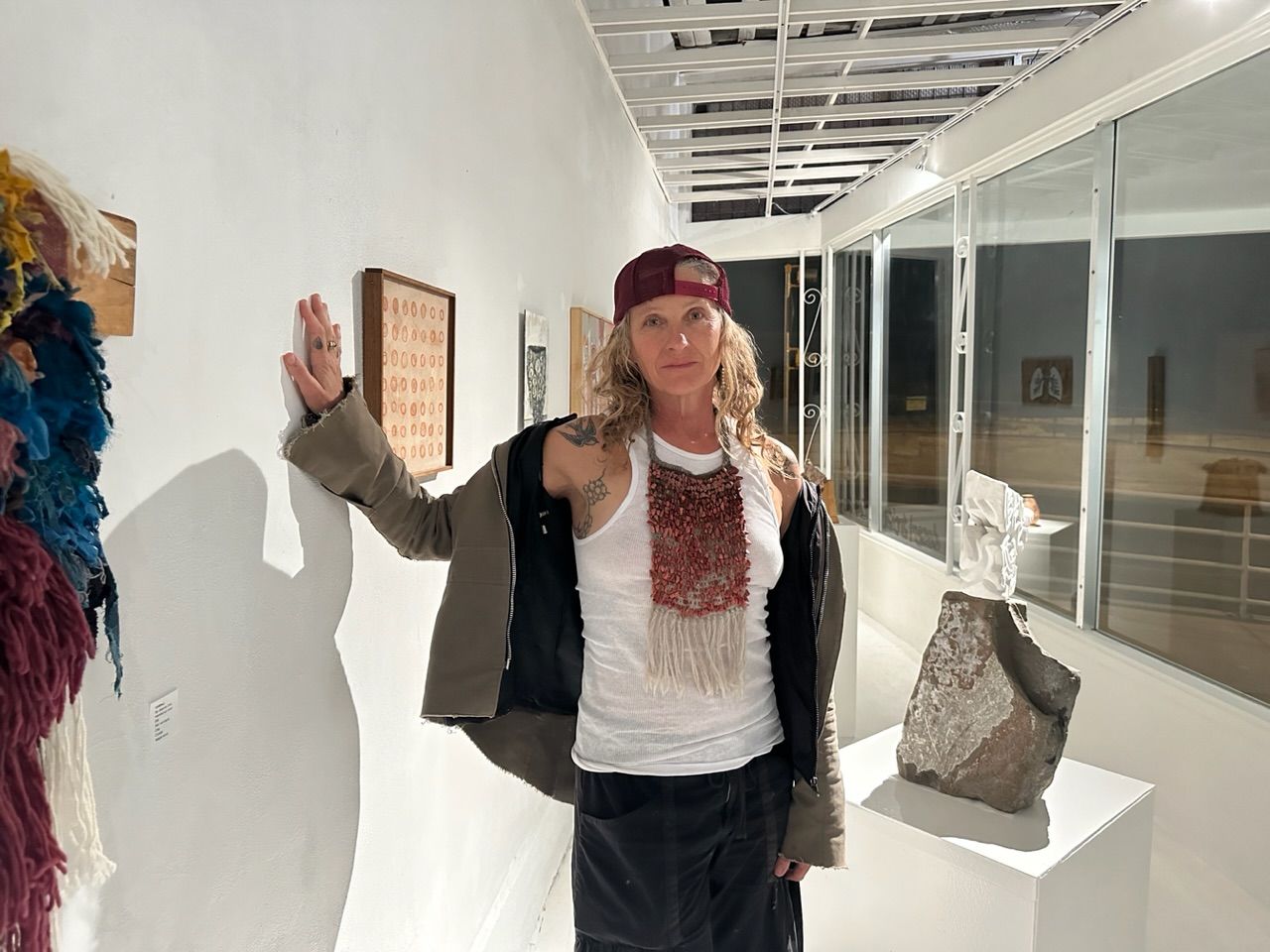 A woman stands in a gallery with white walls wearing a white shirt, red baseball cap, green jacket and a fiber sculpture necklace made of red coral and wool