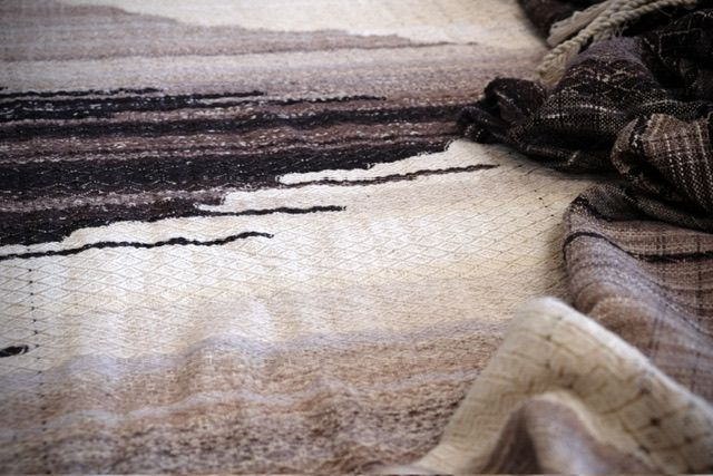 A handwoven length of fabric 30 inches wide and 4.8 meters long in shades of white, gray and black with a textured diamond pattern lays on a wooden floor. 