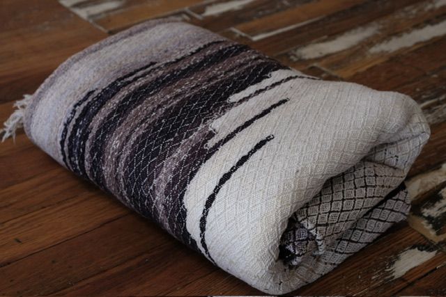 A handwoven length of fabric 30 inches wide and 4.8 meters long in shades of white, gray and black lays on a wooden floor. 