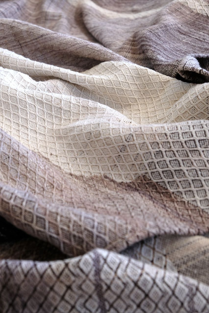 A handwoven length of fabric 30 inches wide and 4.8 meters long in shades of white, gray and black with a textured diamond pattern lays on a wooden floor. 