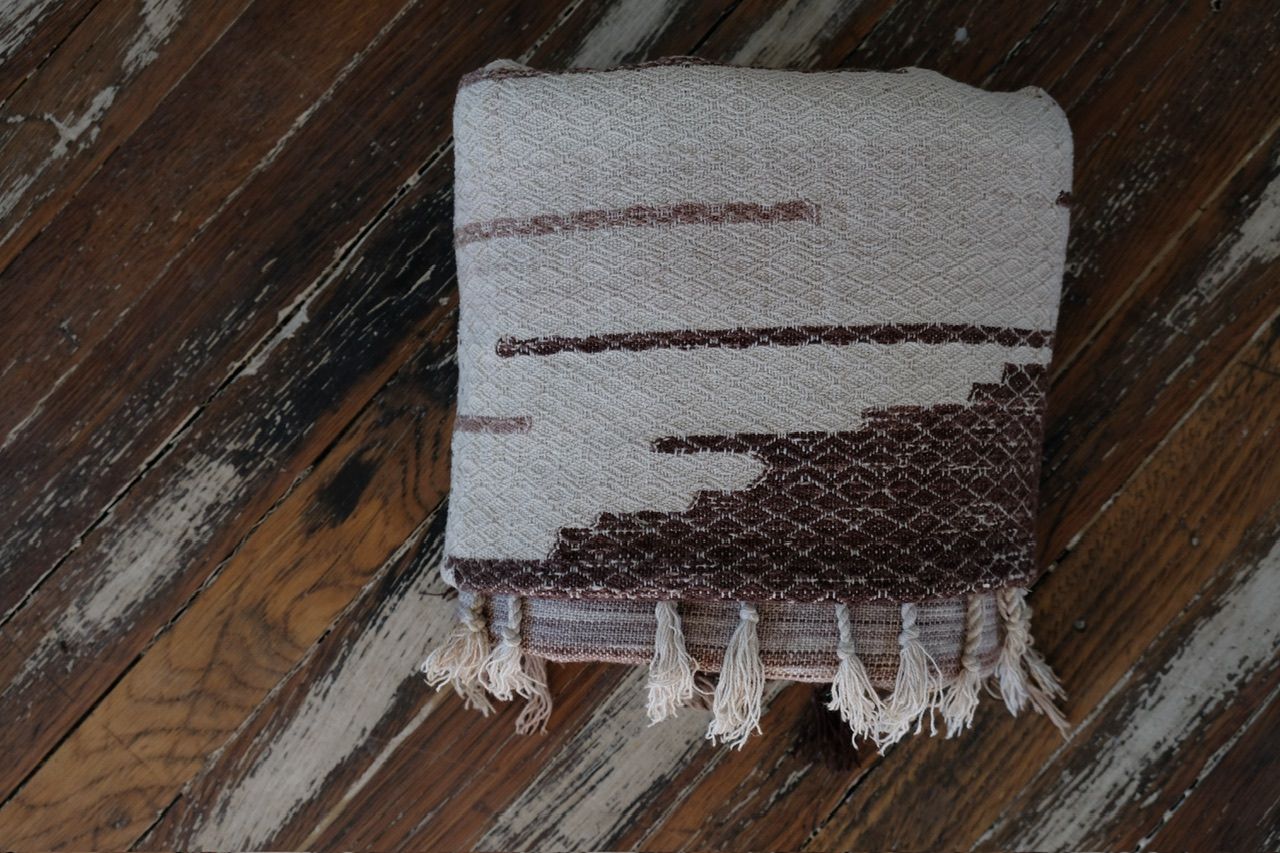 A large piece of handwoven fabric with a peach triangle on one end in shades of brown, cream and white rests on a dark wood floor