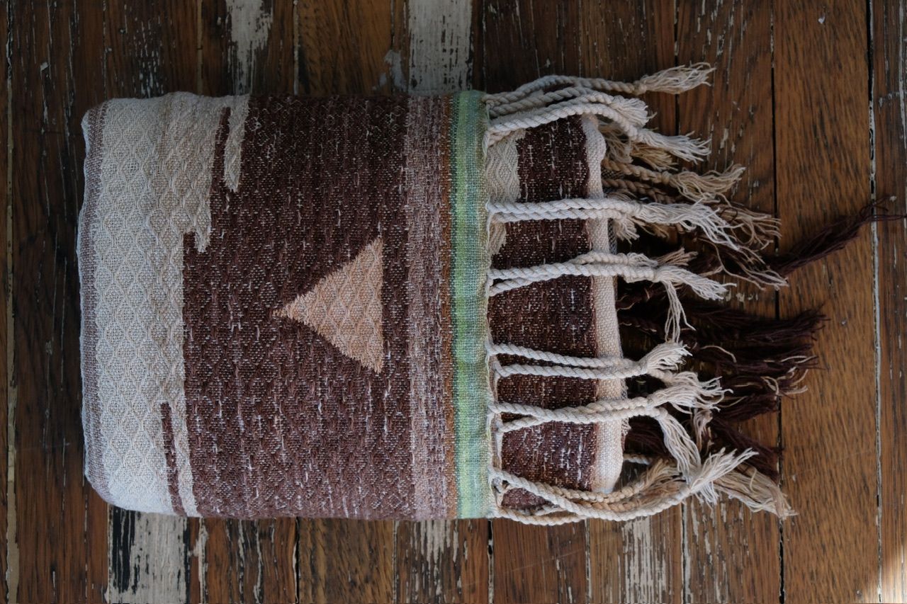 A large piece of handwoven fabric with a peach triangle on one end in shades of brown, cream and white rests on a dark wood floor