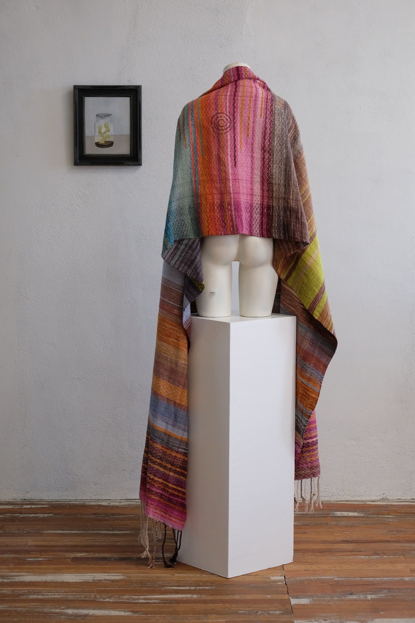 A brightly colored handwoven shawl is draped on a white mannequin in a white gallery space with wood floors.