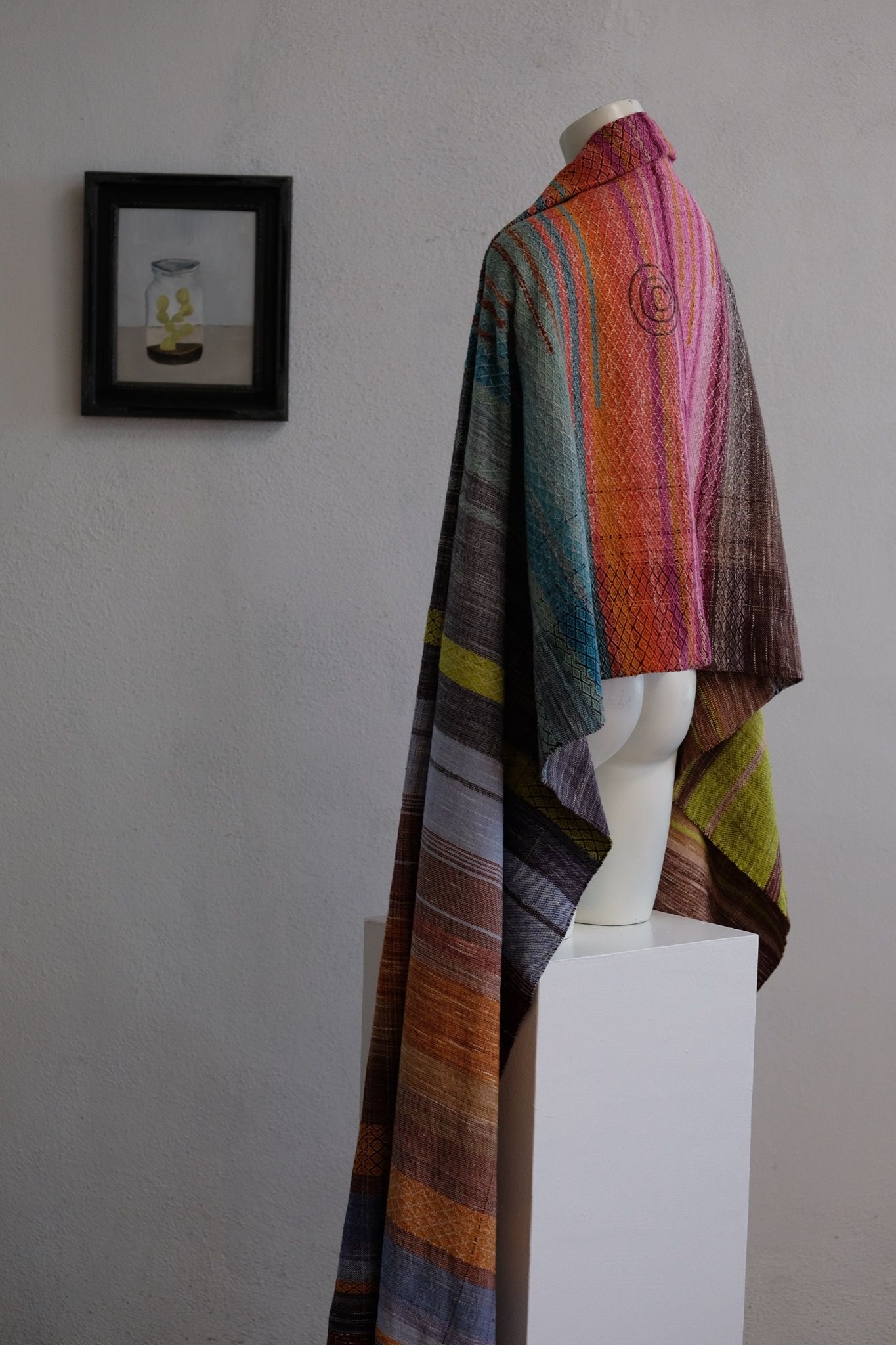A brightly colored handwoven shawl is draped on a white mannequin in a white gallery space with wood floors.
