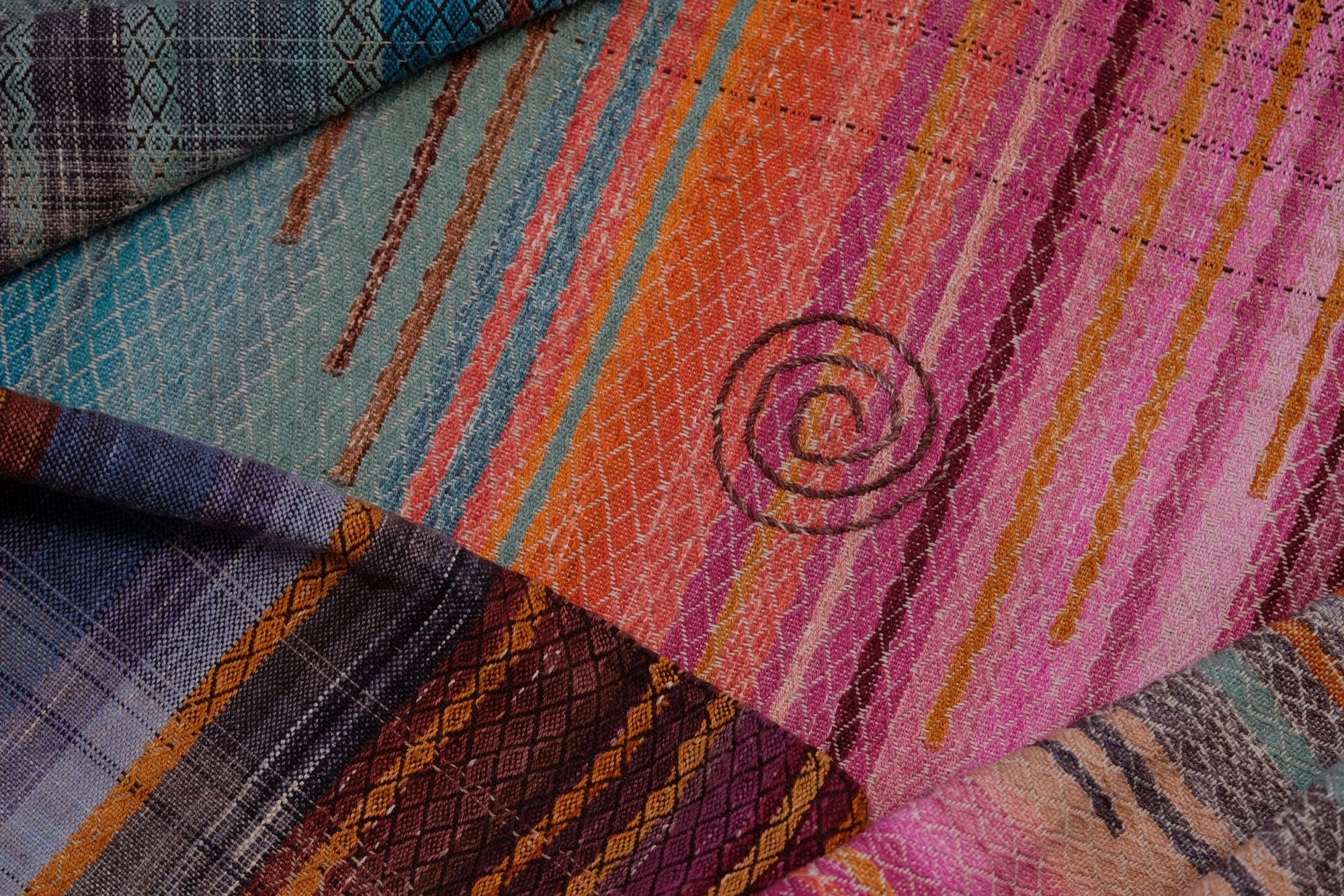 A detailed image of a brightly colored piece of handwoven fabric laying on a wood floor. It is in all shades of sunset pink, orange and gold, as well as blue and green. it has a spiral and crescent moon design on it as well