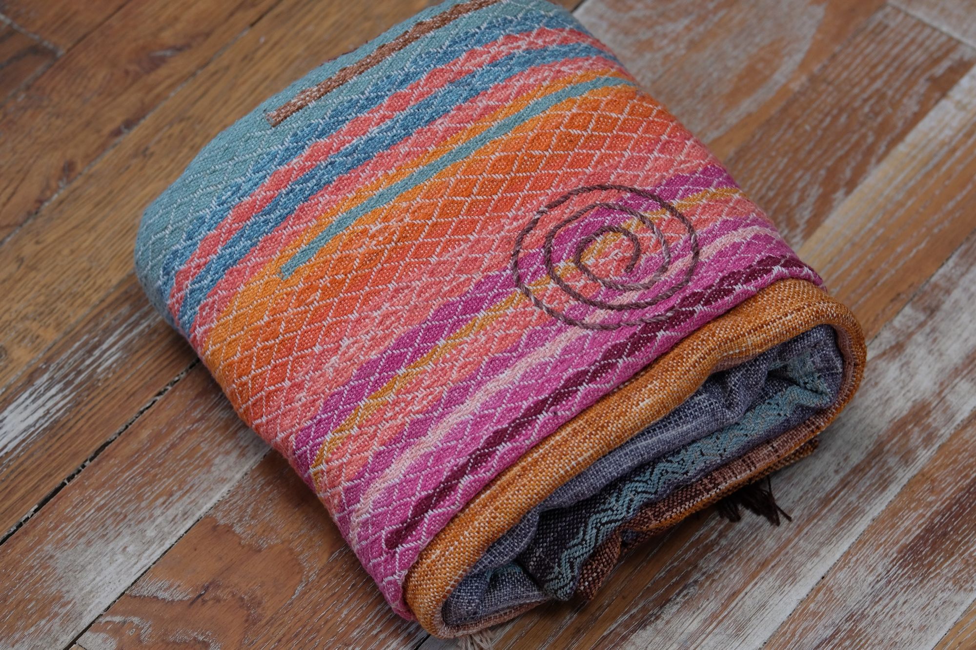 A detailed image of a brightly colored piece of handwoven fabric laying on a wood floor. It is in all shades of sunset pink, orange and gold, as well as blue and green. it has a spiral and crescent moon design on it as well