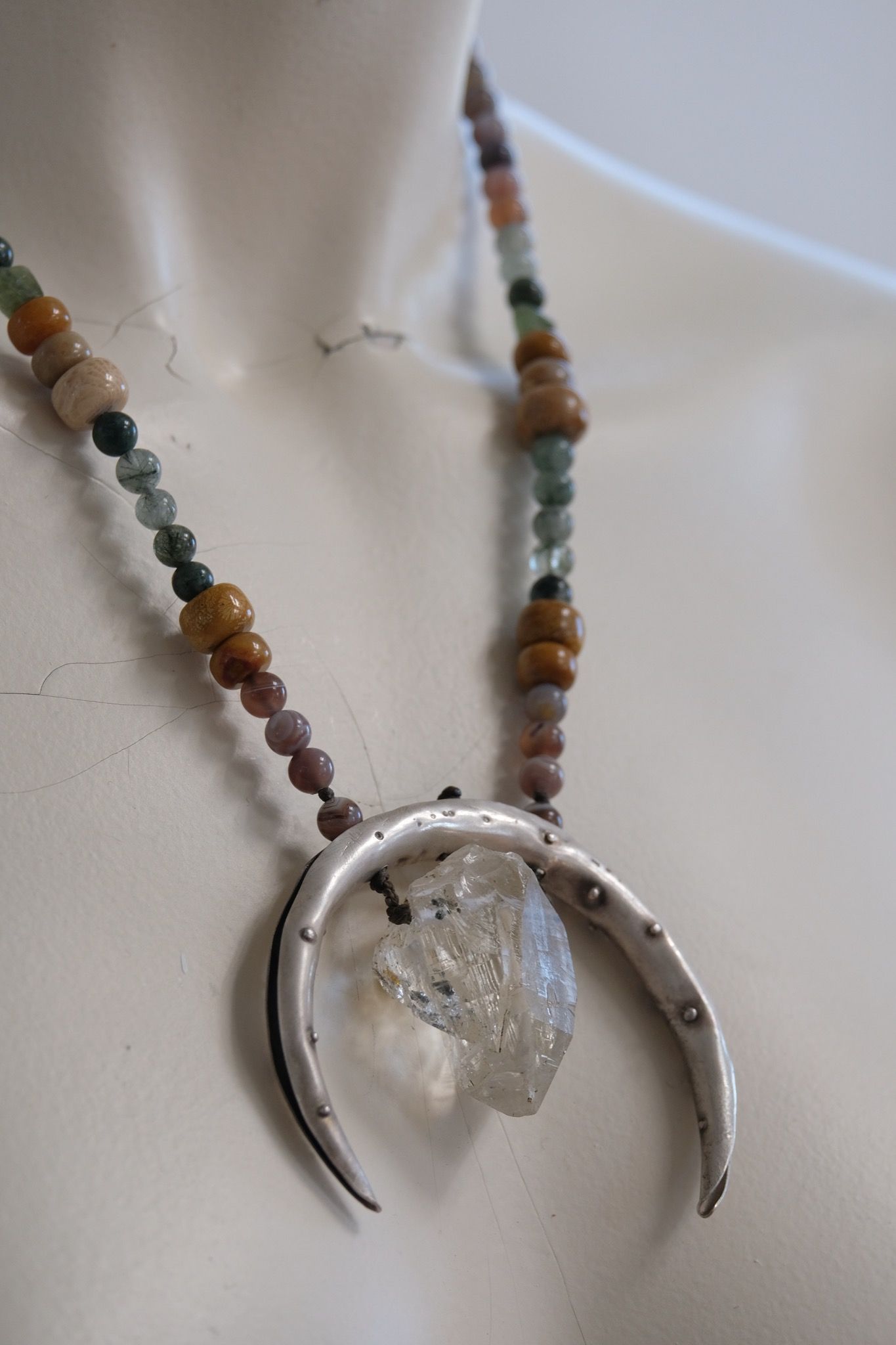 A necklace made of a silver moon, clear quartz crystal, earth-tone and green beads rests on a white mannequin