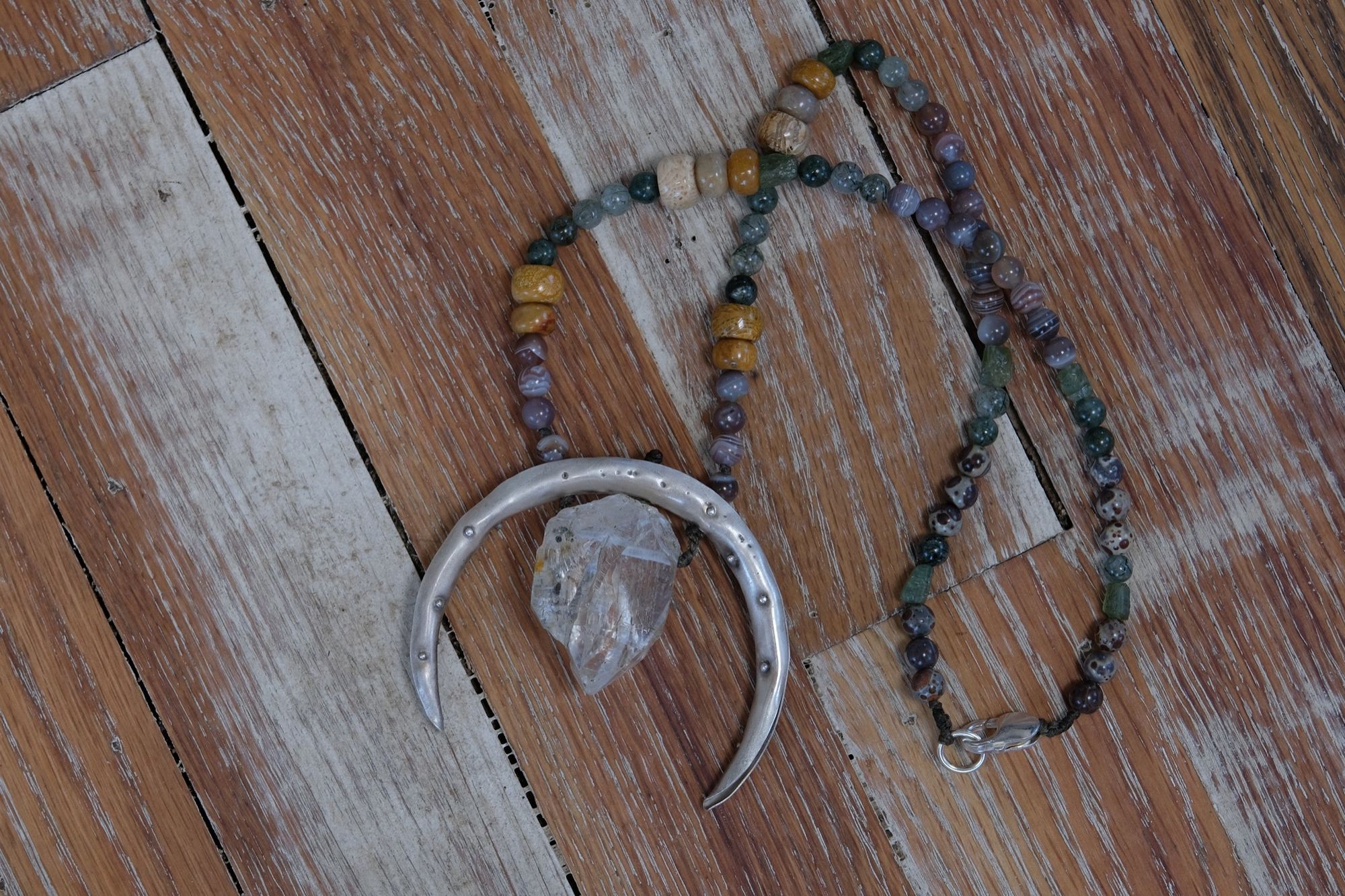 A necklace made of a silver moon, clear quartz crystal, earth-tone and green beads rests on a wood floor