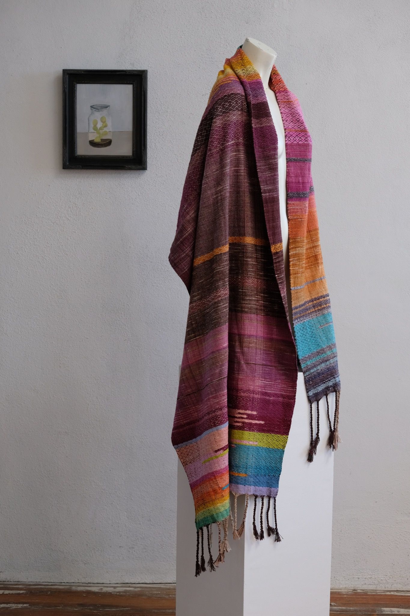 Handwoven fabric with a diamond pattern and vivid sunrise colors of blue, purple, orange, pink and yellow rests on a mannequin in a gallery space with a paining of a cactus in a jar behind it