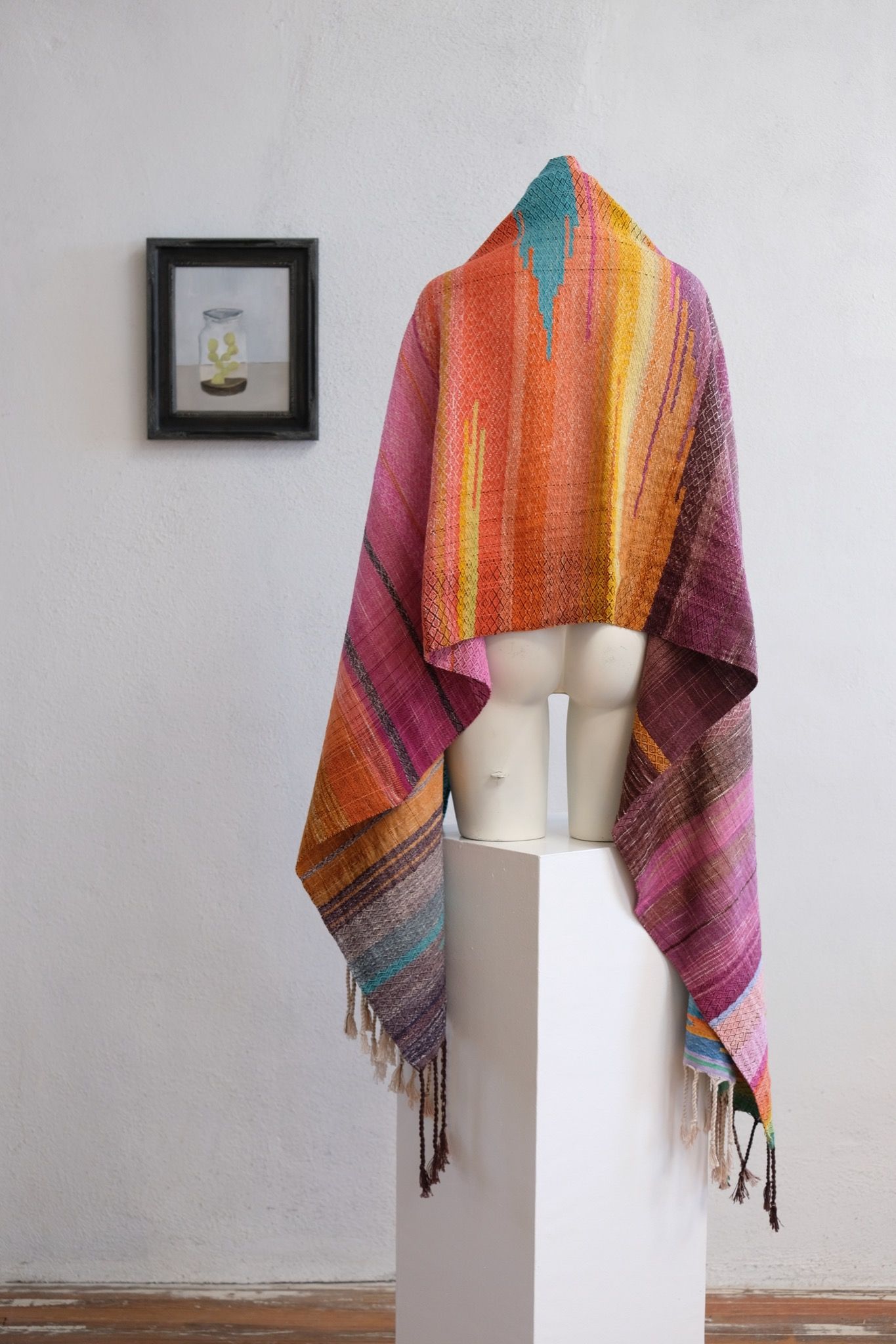 Handwoven fabric with a diamond pattern and vivid sunrise colors of blue, purple, orange, pink and yellow rests on a mannequin in a gallery space with a paining of a cactus in a jar behind it