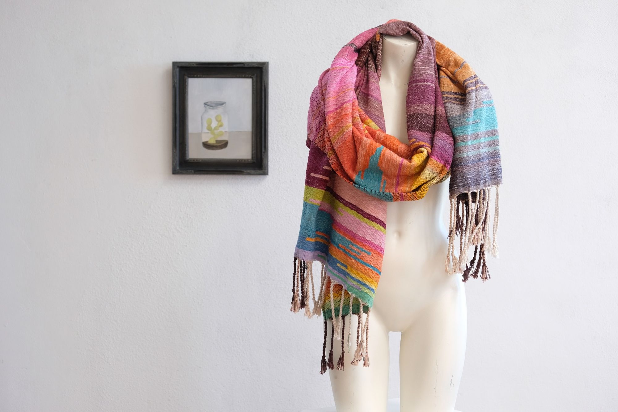 Handwoven fabric with a diamond pattern and vivid sunrise colors of blue, purple, orange, pink and yellow rests on a mannequin in a gallery space with a paining of a cactus in a jar behind it