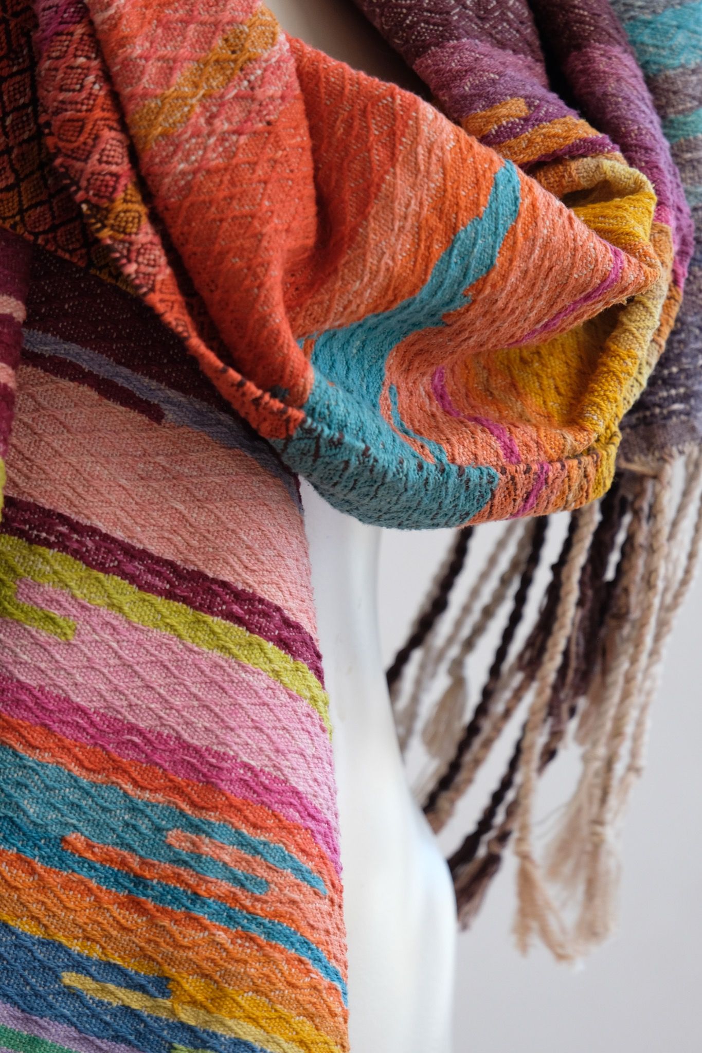 Handwoven fabric with a diamond pattern and vivid sunrise colors of blue, purple, orange, pink and yellow rests on a mannequin in a gallery space with a paining of a cactus in a jar behind it