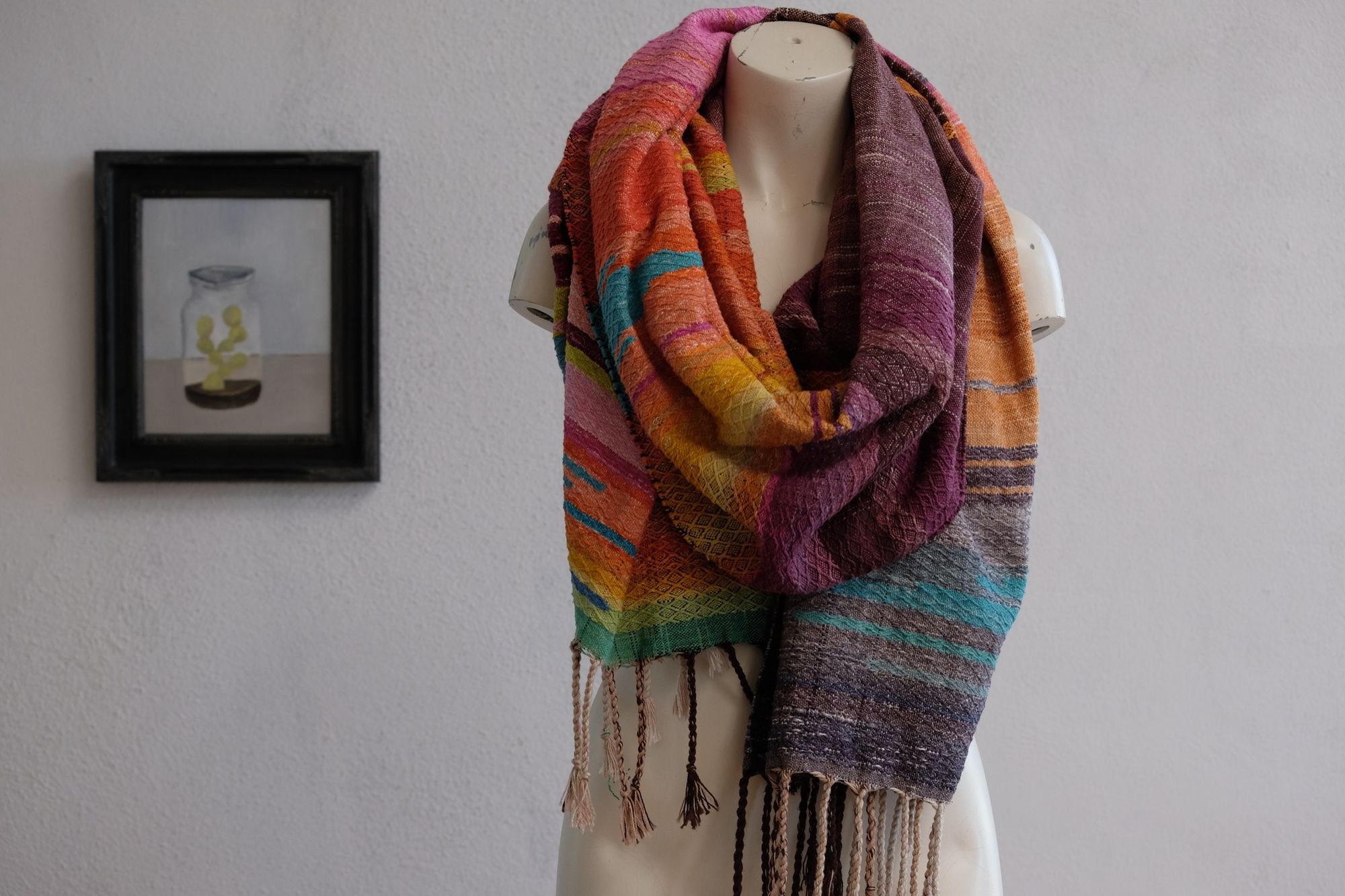 Handwoven fabric with a diamond pattern and vivid sunrise colors of blue, purple, orange, pink and yellow rests on a mannequin in a gallery space with a paining of a cactus in a jar behind it