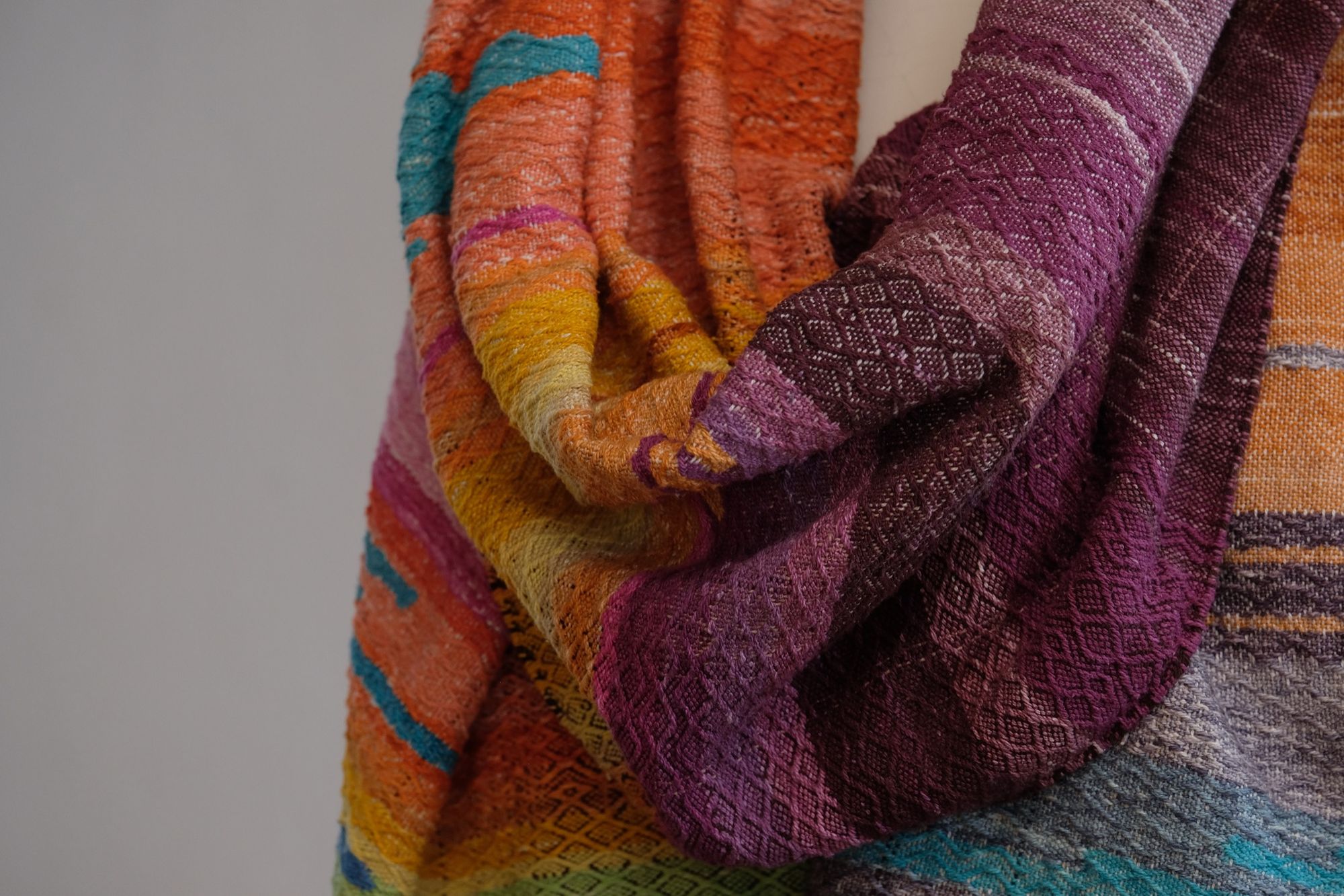 Handwoven fabric with a diamond pattern and vivid sunrise colors of blue, purple, orange, pink and yellow rests on a mannequin in a gallery space with a paining of a cactus in a jar behind it