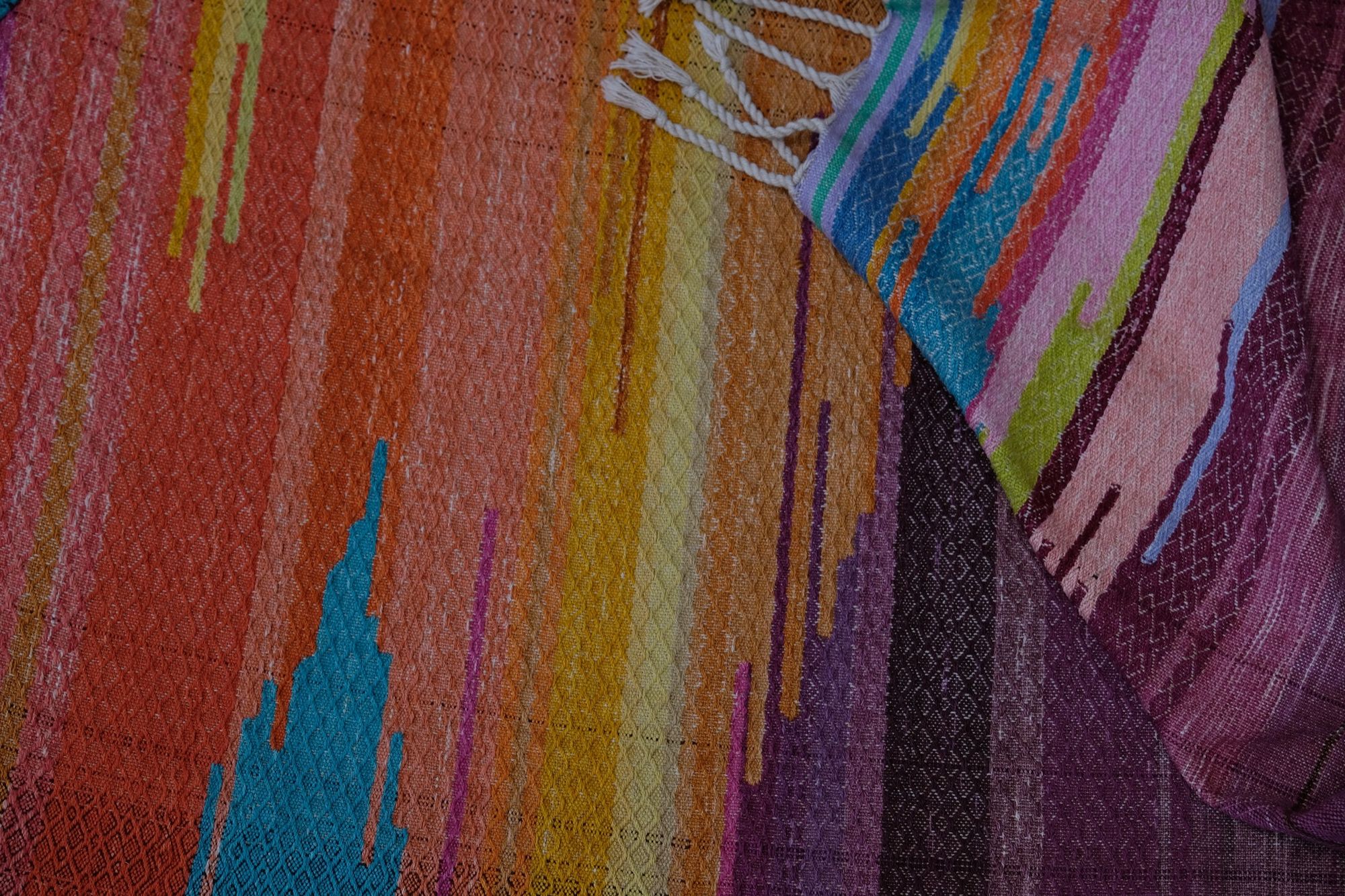 Handwoven fabric with a diamond pattern and vivid sunrise colors of blue, purple, orange, pink and yellow lays on a wooden floor