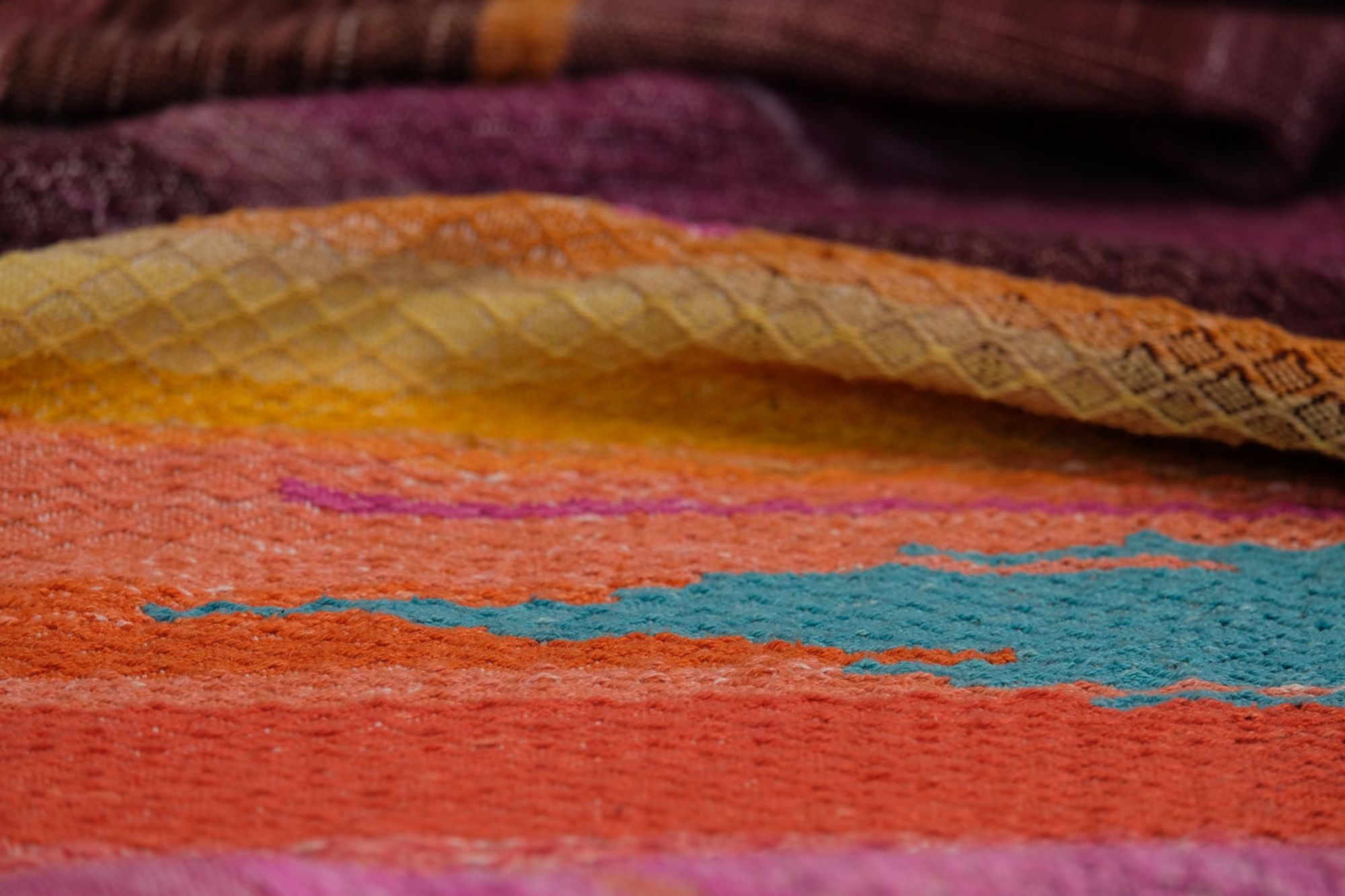 Handwoven fabric with a diamond pattern and vivid sunrise colors of blue, purple, orange, pink and yellow lays on a wooden floor