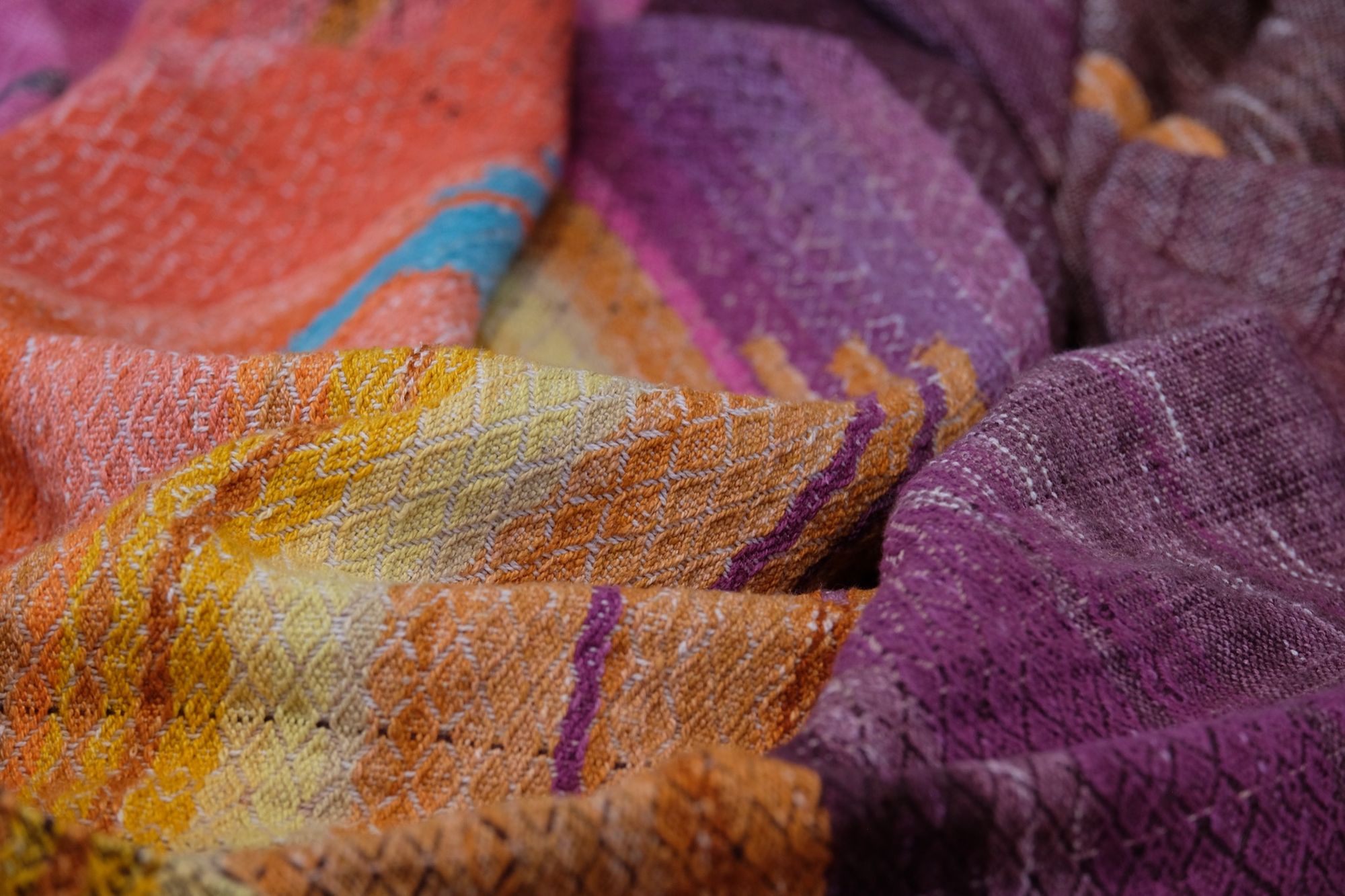 Handwoven fabric with a diamond pattern and vivid sunrise colors of blue, purple, orange, pink and yellow lays on a wooden floor