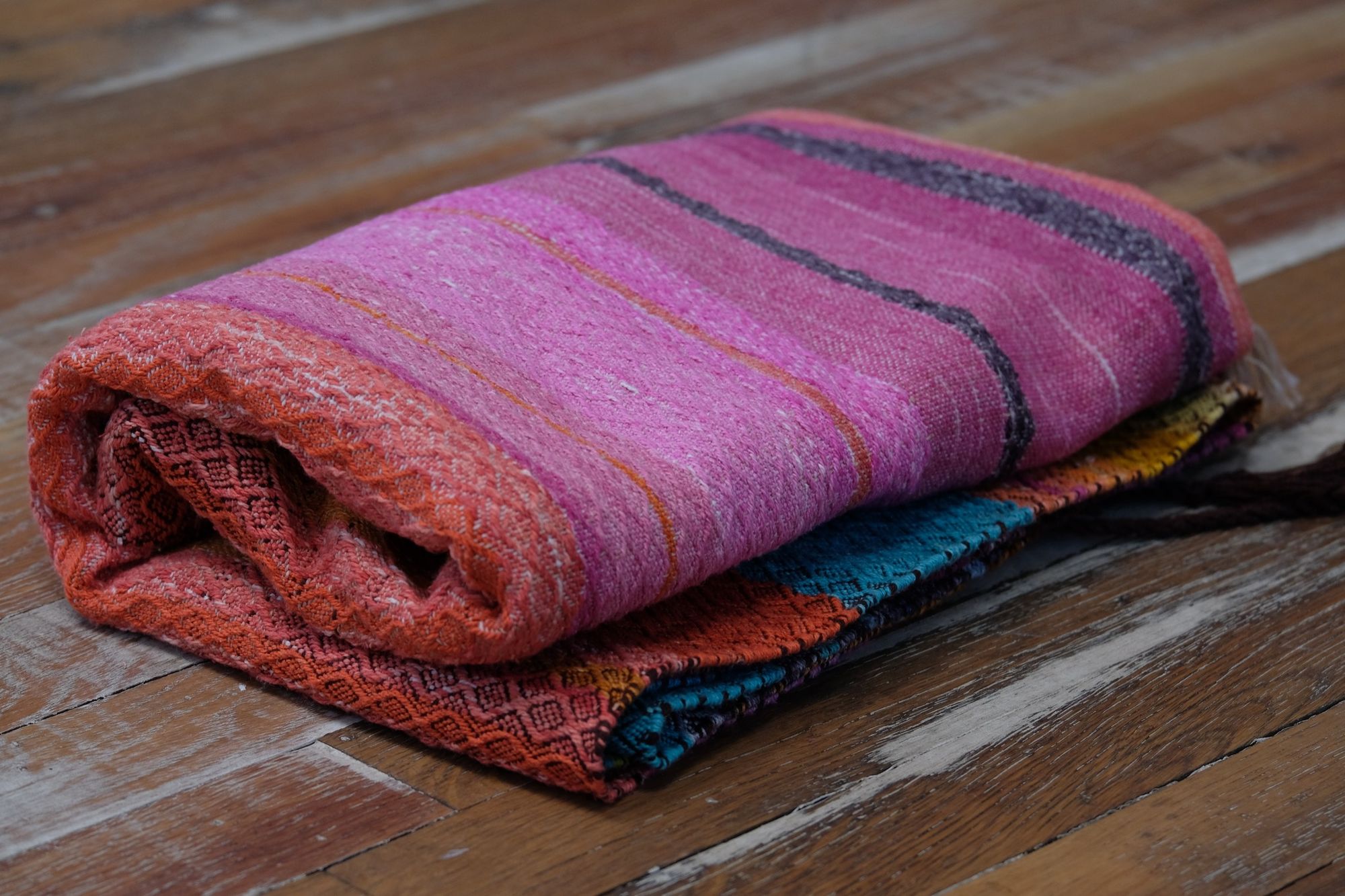 Handwoven fabric with a diamond pattern and vivid sunrise colors of blue, purple, orange, pink and yellow lays on a wooden floor