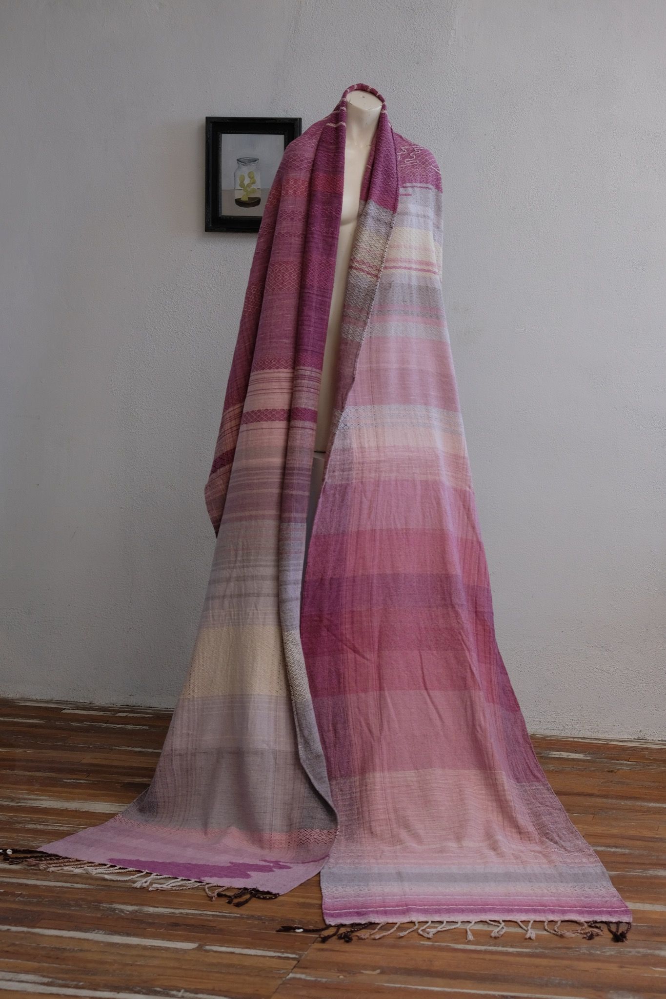 Handwoven fabric lays on a white mannequin in a gallery space. The fabric is naturally dyed with cochineal in all shades of pink and fuchsia with a design of a river and three moons on it