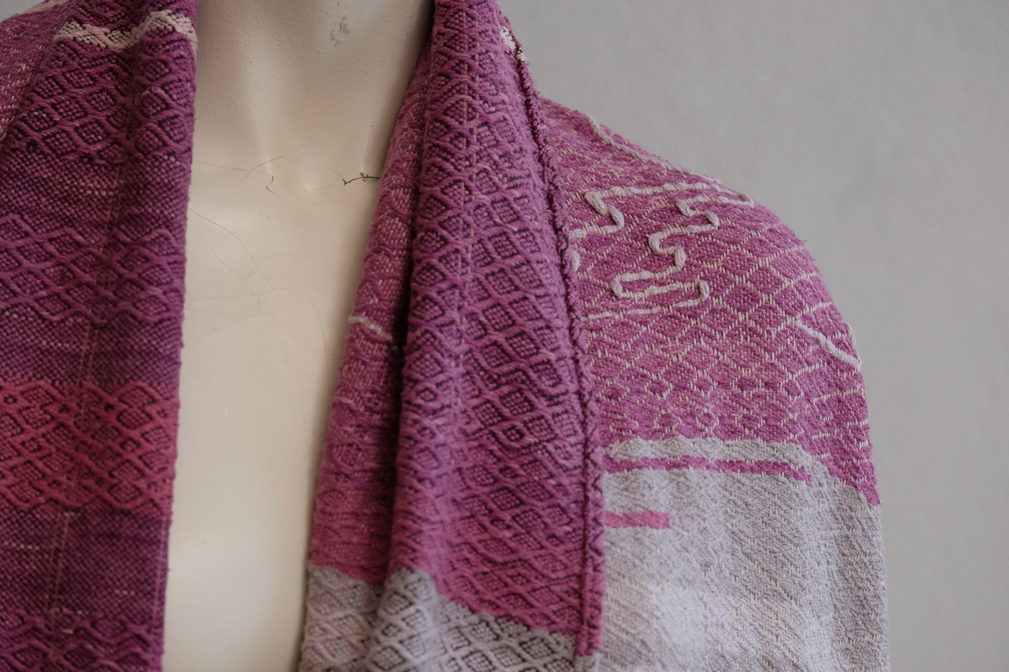 Handwoven fabric lays on a white mannequin in a gallery space. The fabric is naturally dyed with cochineal in all shades of pink and fuchsia with a design of a river and three moons on it