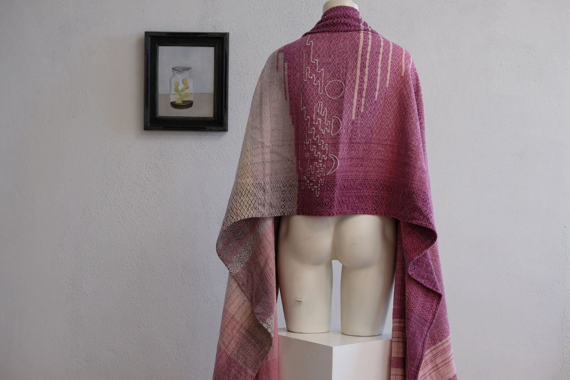 Handwoven fabric lays on a white mannequin in a gallery space. The fabric is naturally dyed with cochineal in all shades of pink and fuchsia with a design of a river and three moons on itHandwoven fabric lays on a wooden floor in a gallery space. The fabric is naturally dyed with cochineal in all shades of pink and fuchsia with a design of a river and three moons on it
