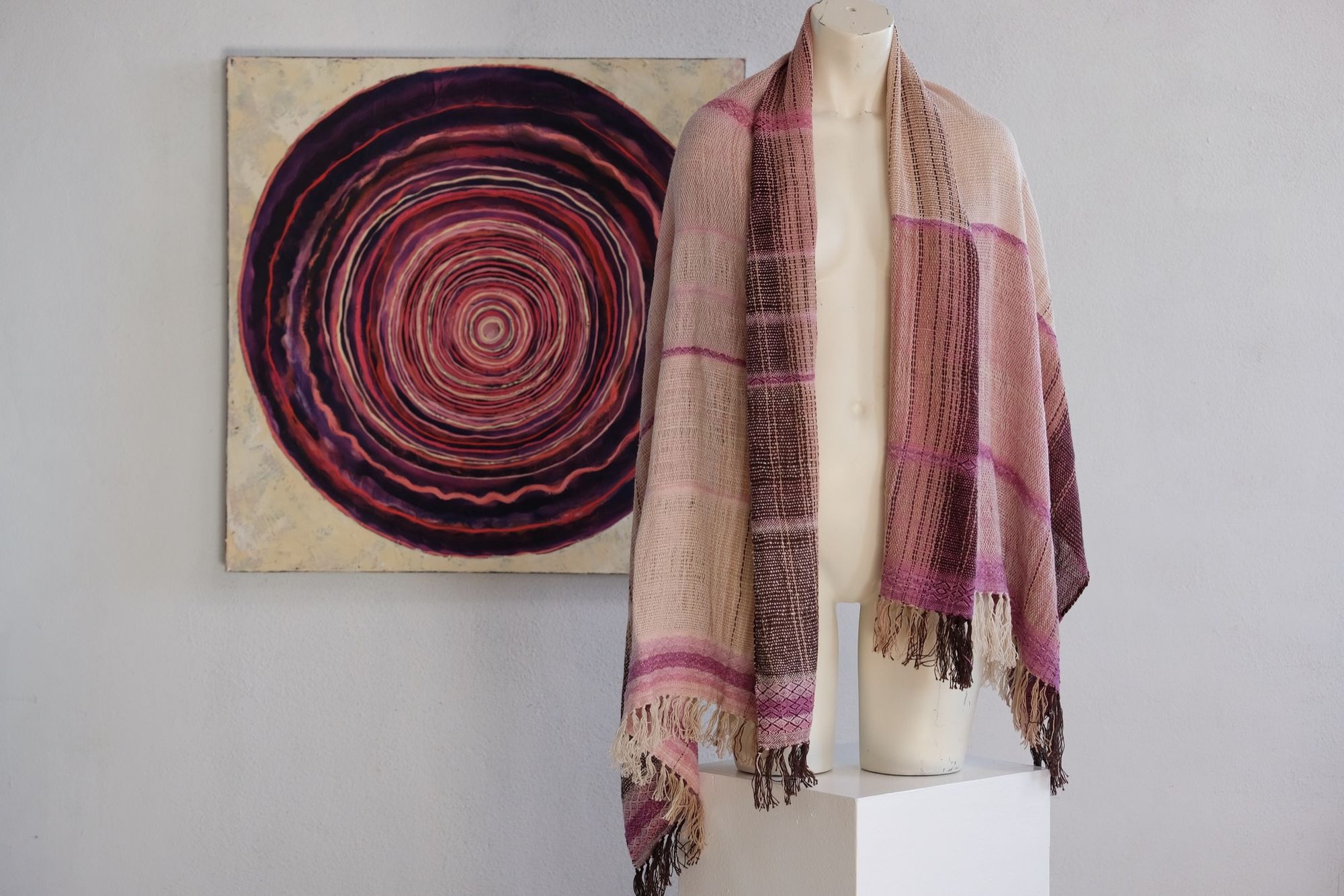 Handwoven scarf naturally dyed shades of pink and fuchsia on a white mannequin in a white gallery space in the background is an oil painting of a purple vortex-like shape