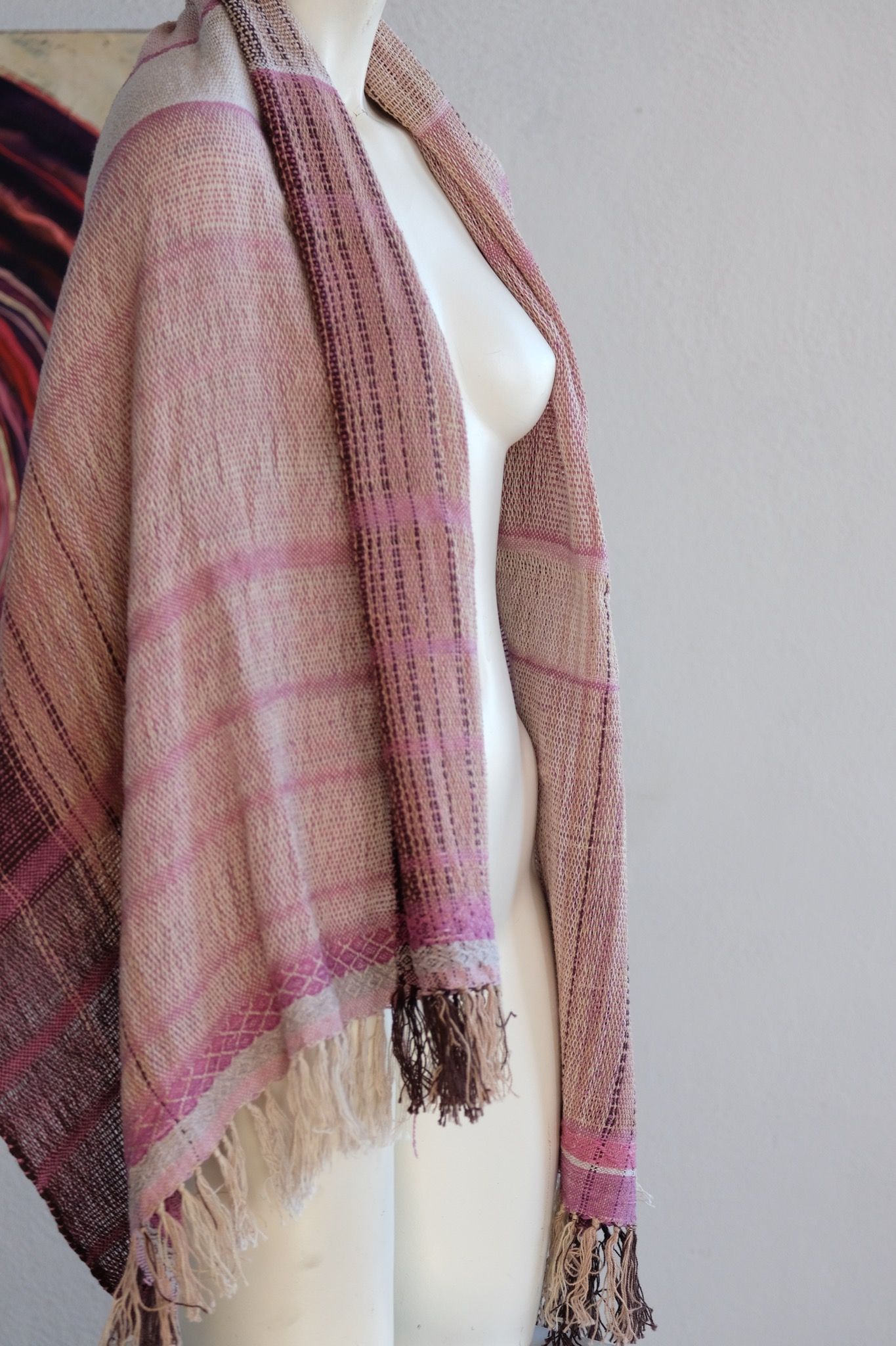 Handwoven scarf naturally dyed shades of pink and fuchsia on a white mannequin in a white gallery space in the background is a large oil painting which depicts a purple vortex like shape