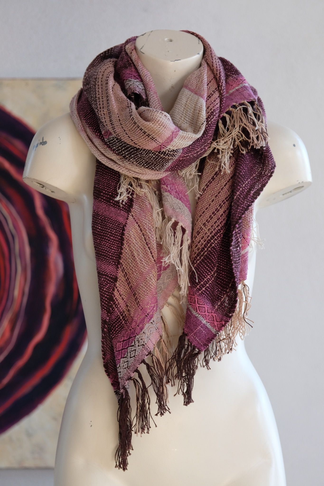 Handwoven scarf naturally dyed shades of pink and fuchsia on a white mannequin in a white gallery space in the background is a large oil painting which depicts a purple vortex like shape