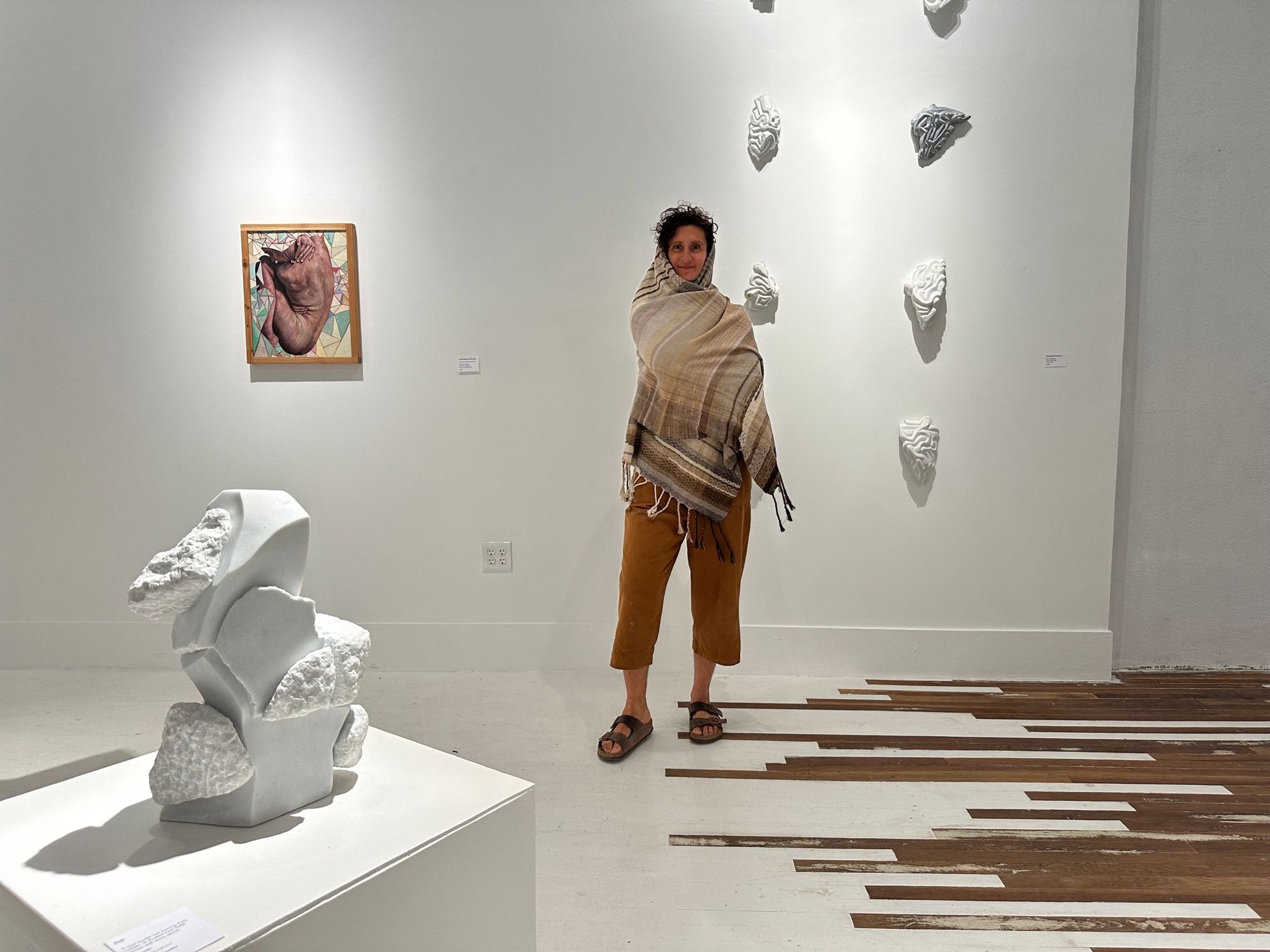A woman wearing ochre colored overalls wears a brown, tan, white gradation shawl while moving around in a white and wooden gallery space with marble sculptures and oil paintings all around