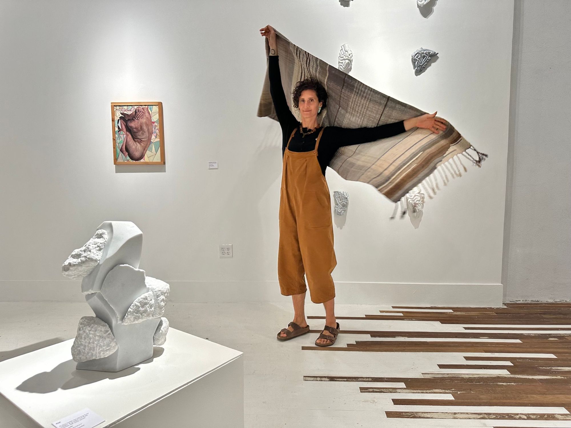 A woman wearing ochre colored overalls wears a brown, tan, white gradation shawl while moving around in a white and wooden gallery space with marble sculptures and oil paintings all around