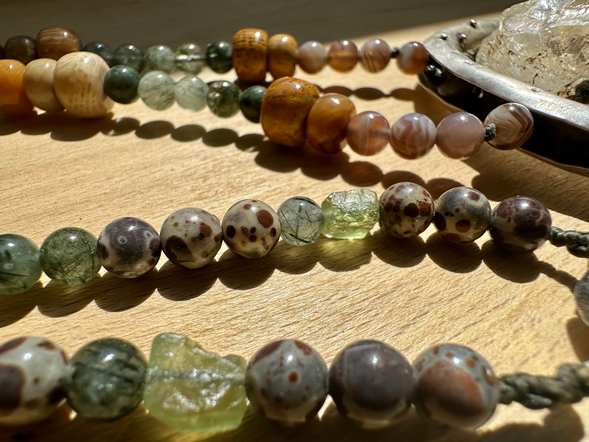 A necklace made of a silver moon, clear quartz crystal, earth-tone and green beads rests on a wood floor