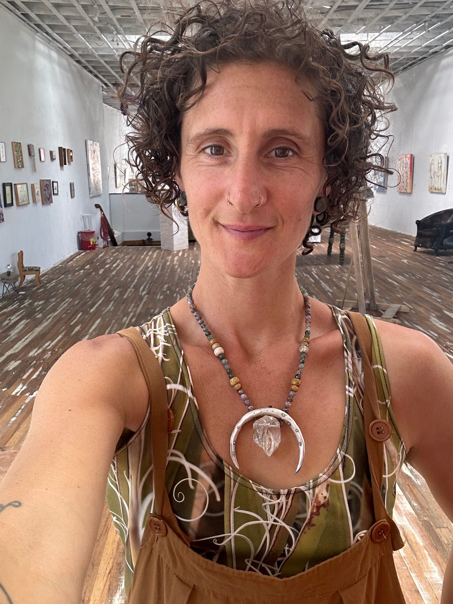 a woman in a large gallery space, wearing tan overalls wears A necklace made of a silver moon, clear quartz crystal, earth-tone and green beads 