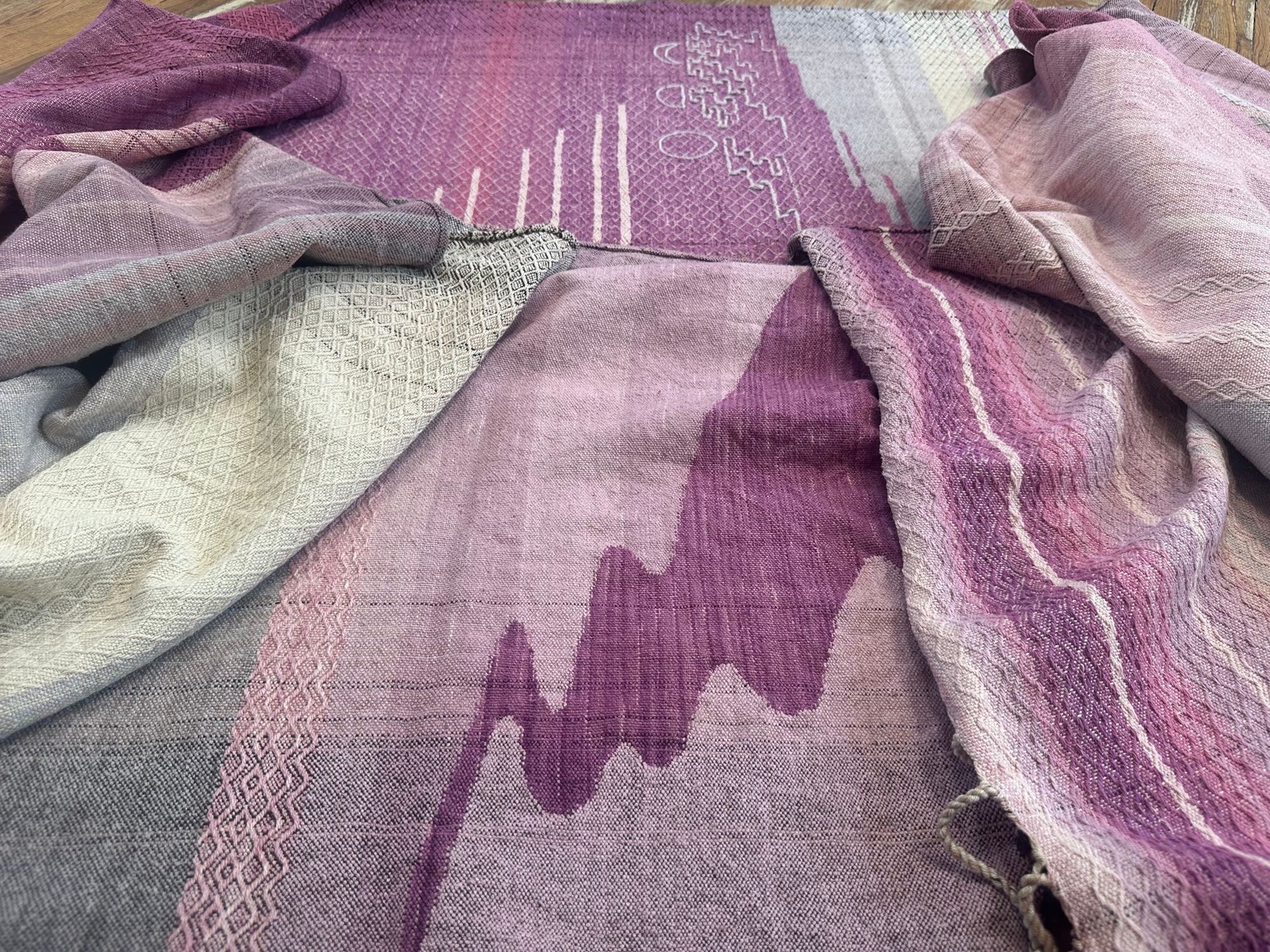Handwoven fabric lays on a wooden floor in a gallery space. The fabric is naturally dyed with cochineal in all shades of pink and fuchsia with a design of a river and three moons on it