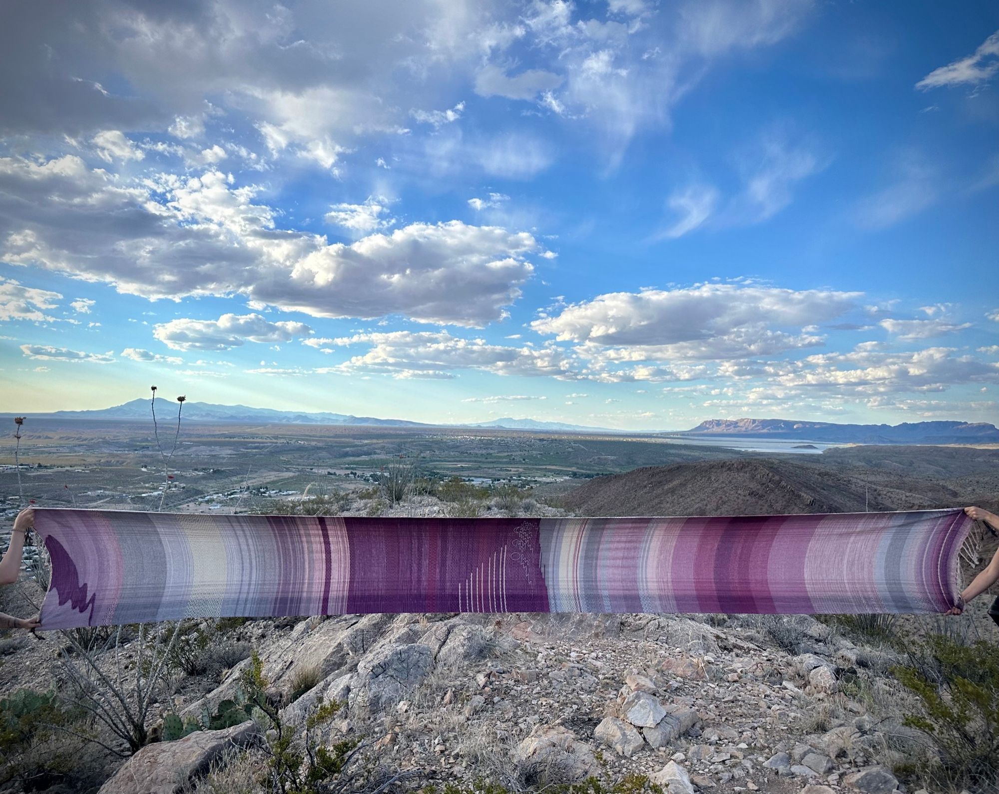 four hands hold out a piece of handwoven fabric that is naturally dyed with cochineal in all shades of pink and fuchsia against a mountain desert landscape