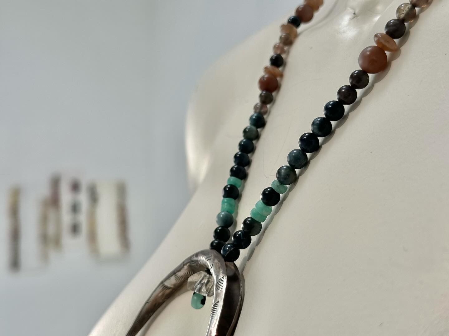 A white mannequin wears a necklace with a silver crescent, moon, emerald, and cats eye beads in a gallery space