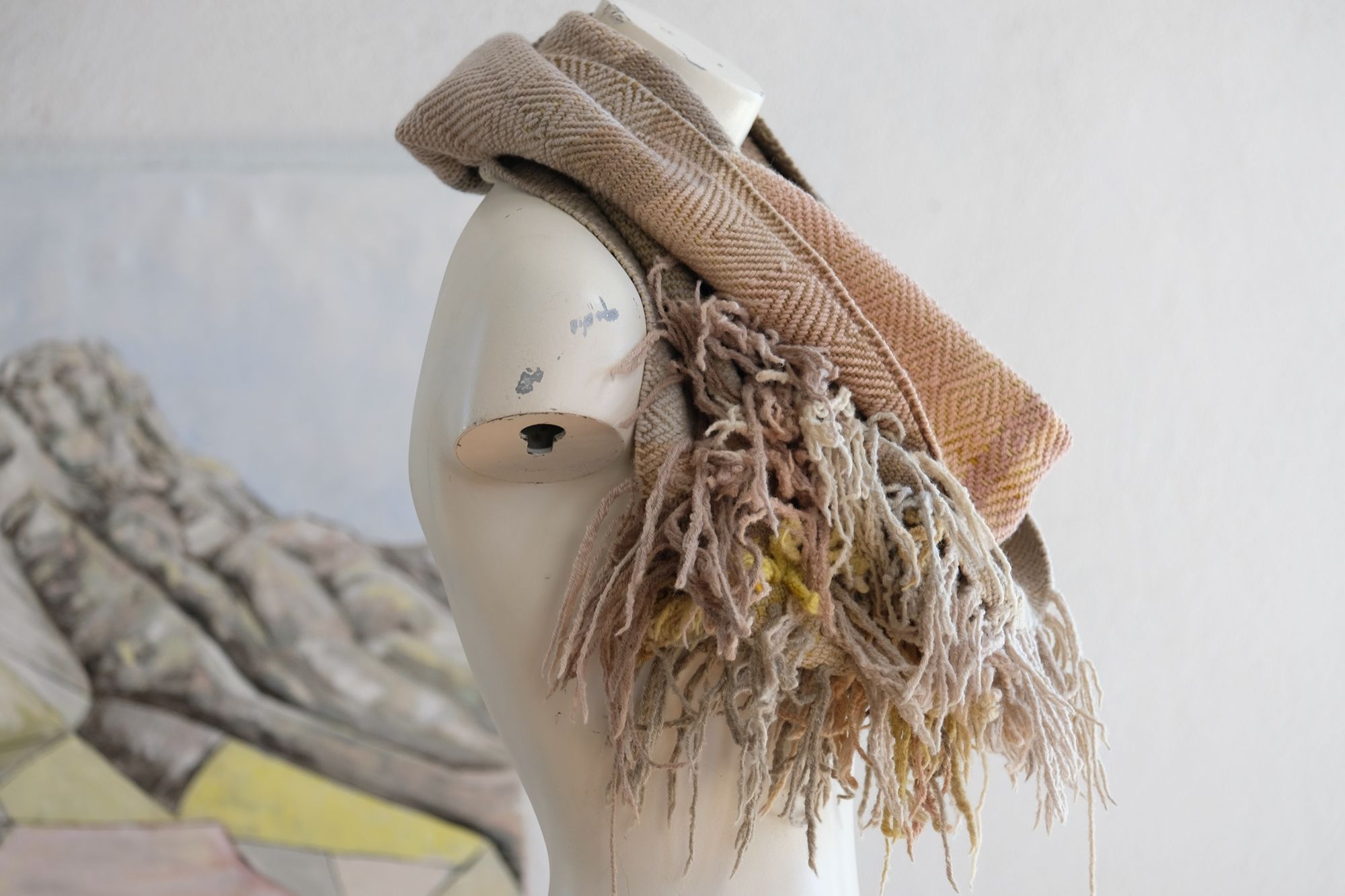 A white painted mannequin torso wears a handwoven naturally dyed highly textured and fringed loop scarf in soft shades of pink, grey, yellow and tan