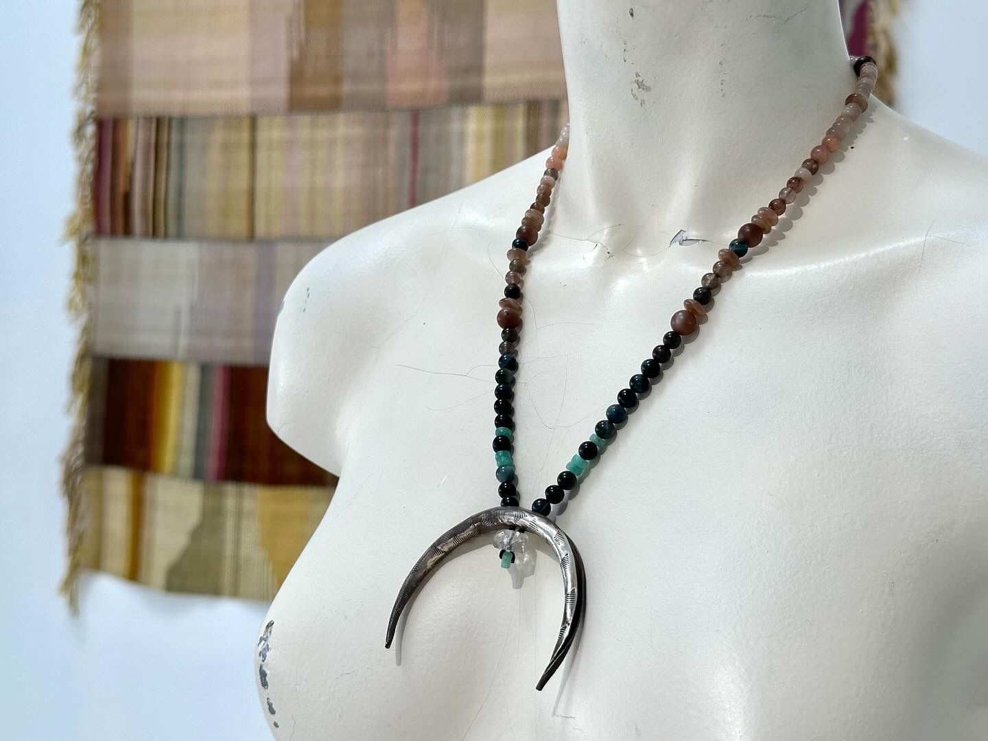 A white mannequin wears a necklace with a silver crescent, moon, emerald, and cats eye beads in a gallery space. Behind it is a large handwoven naturally died blanket.