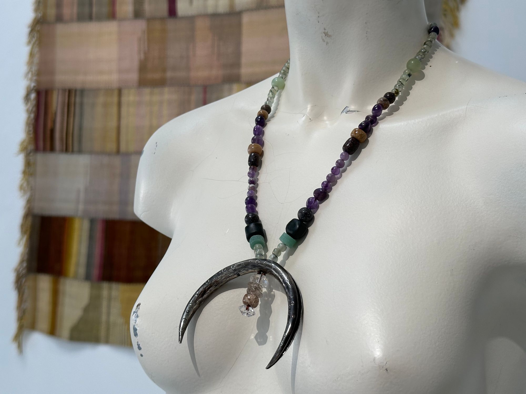 A white mannequin wears a necklace with a silver crescent, moon and green and purple, semi precious stone beads Of fossilized dinosaur bone, coral, Afghan Jade, peridot, Amethyst