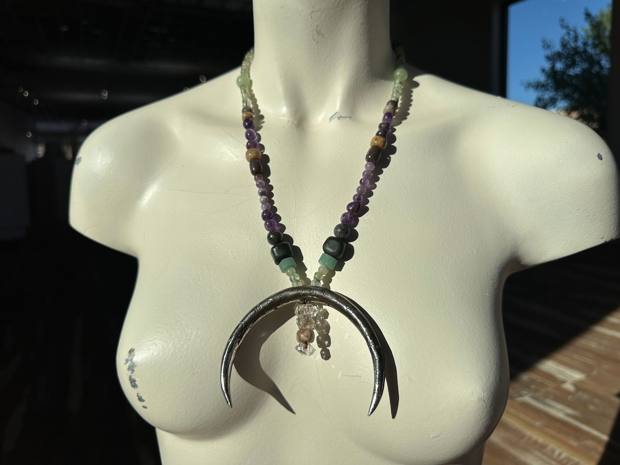 A white mannequin wears a necklace with a silver crescent, moon and green and purple, semi precious stone beads