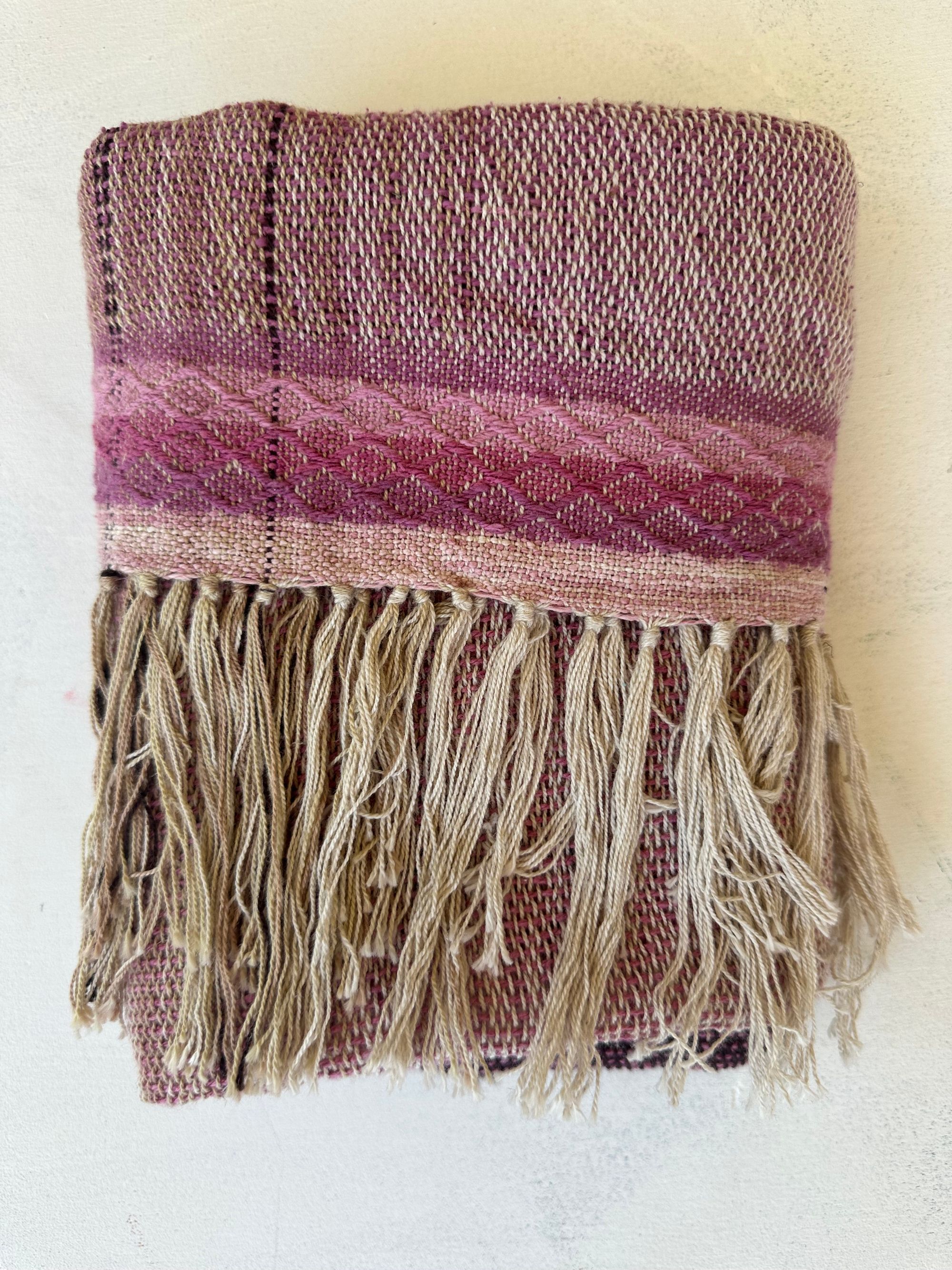 A folded piece of cloth rests on a white floor. The cloth is handwoven out of organic cotton linen and cochineal dyed raw Silk